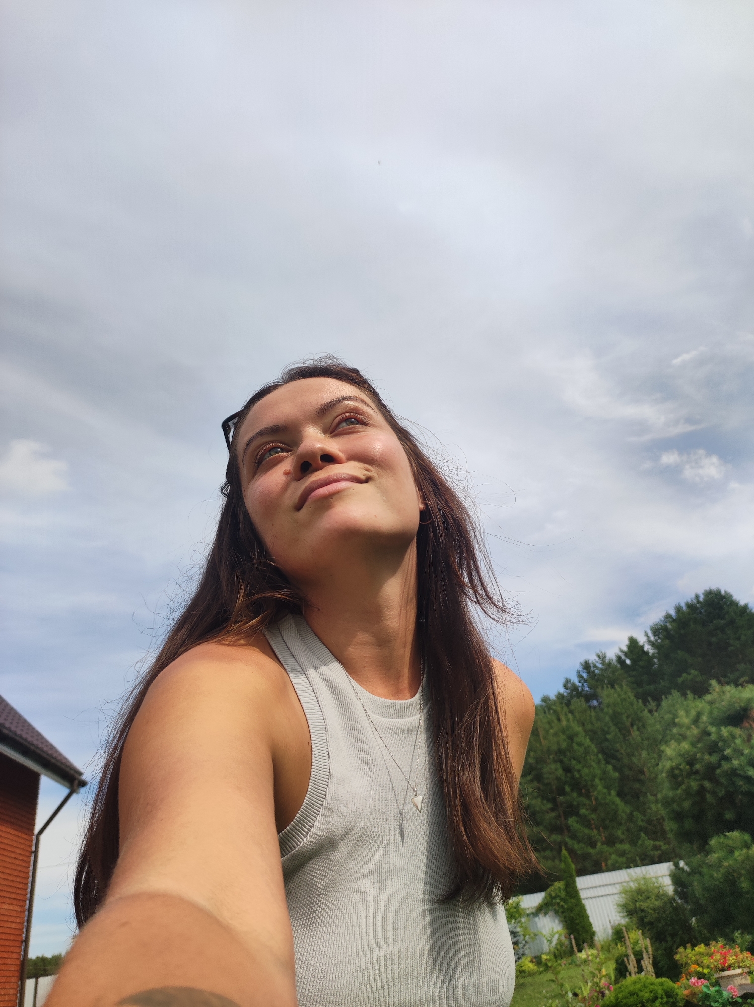 In this medium shot photograph, a young woman with long, dark brown hair looks up towards a cloudy sky. She is wearing a light grey, ribbed tank top and a silver necklace with a heart-shaped pendant. Her face is illuminated by sunlight, casting highlights on her skin. She has sunglasses pushed up on her head and a subtle smile on her lips. The background features a lush green treeline with tall pine trees and a glimpse of a red-tiled roof of a building on the left. A white fence and a manicured garden with various plants and flowers are visible in the mid-ground. The image appears to be taken outdoors, likely in a residential area or backyard, possibly in the city of Fokino, Russia.
