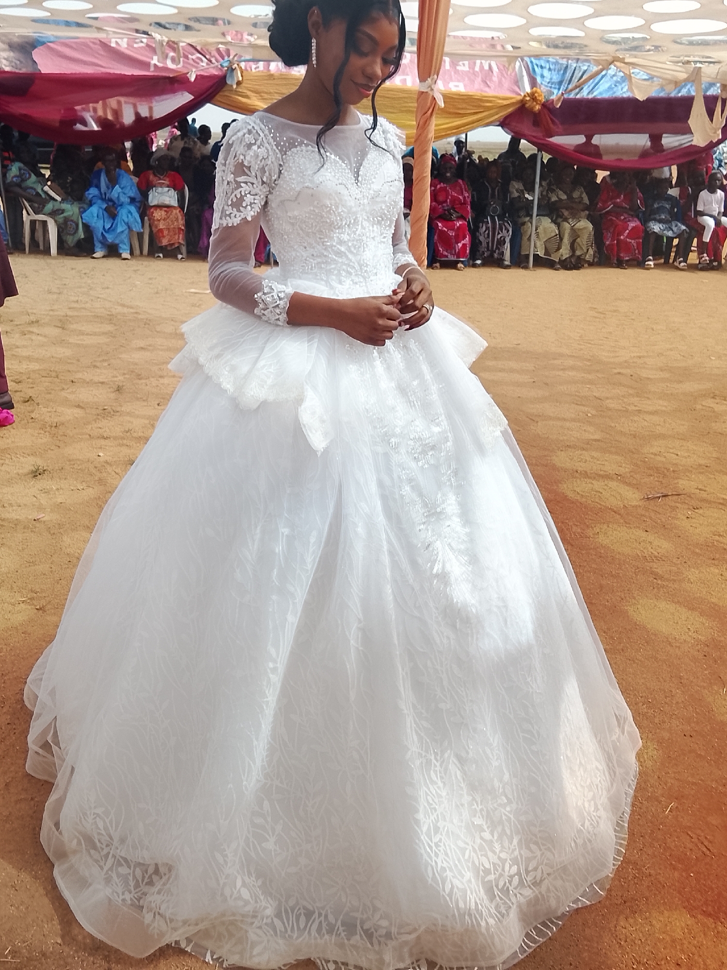 A young woman in a white wedding dress stands on reddish-brown ground in Dogon Dinia, Nigeria. The dress has a sheer, lace-sleeved bodice with a sweetheart neckline and a full, layered skirt. Behind her, under a canopy draped with maroon and gold fabric, are seated people dressed in colorful clothing. The ground is bare, suggesting an outdoor setting.