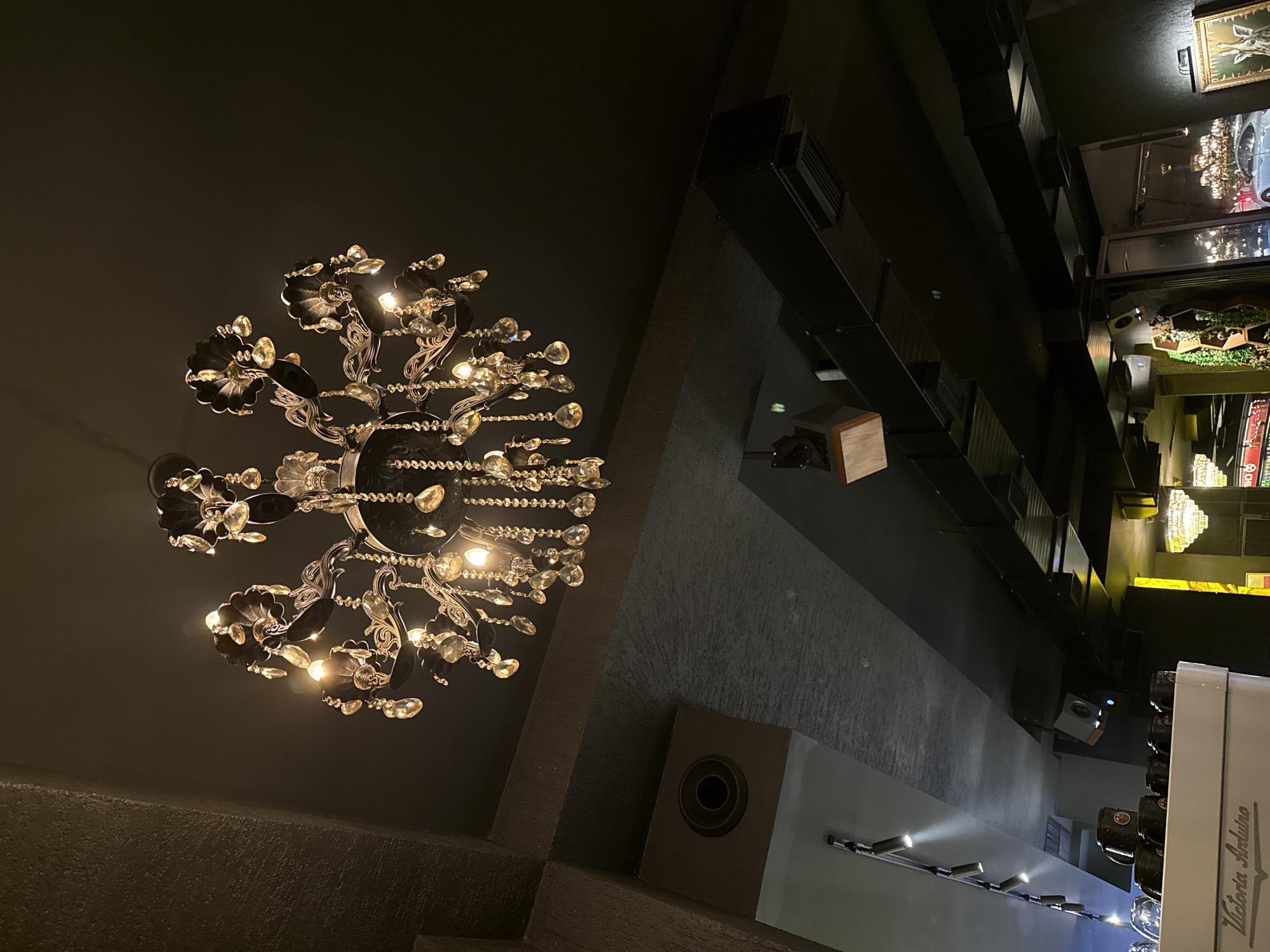 The image shows the interior of a business in Unknown, Unknown. The low-angle view highlights a large, ornate chandelier hanging from the ceiling, which is a dark, muted color. The walls are textured and a dark grey color. The lighting is dim, and the scene appears to be inside a shop or store, indicated by shelving units stocked with merchandise. A section of a white machine with text "Victoria Arduino" on it is visible in the bottom right corner. There is also a series of spotlights along the bottom edge of the image.