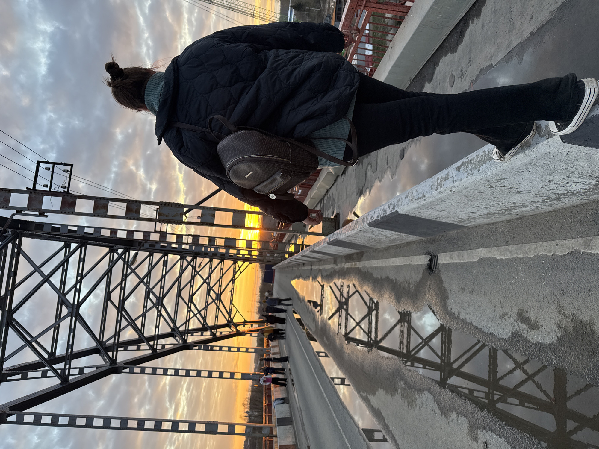 This media file captures a serene moment on a bridge in Dmitrov, Russia, during what appears to be either sunset or sunrise. The image is taken from behind and slightly below a person standing on the bridge, looking out towards the vibrant horizon.
The main subject is seen from the back, wearing a black quilted jacket, a light blue-green turtleneck sweater, black trousers, and black sneakers with white soles. Their dark hair is pulled up into a bun. They are carrying a small, dark brown backpack adorned with a repeating "PDPDPD..." pattern and an unidentifiable metallic brand logo. The person is positioned next to a concrete barrier that forms the edge of the bridge's pathway.
To the left of the frame, a large metal truss bridge structure is prominently featured, its geometric framework silhouetted against the sky. On the right, the concrete surface of the bridge pathway is visible, marked by several puddles. These puddles create captivating reflections, mirroring the detailed truss structure of the bridge and the dramatic sky above.
The sky exhibits a transition from soft grey and white clouds at the top to striking orange and golden hues near the horizon, where the sun's glow is most intense. In the distance, further along the bridge, several indistinct figures of other people can be observed, either walking or also pausing to take in the view. The presence of puddles on the bridge suggests recent rainfall or melting snow. The overall atmosphere is tranquil and contemplative, highlighting the interplay between the industrial architecture of the bridge and the natural beauty of the sky.