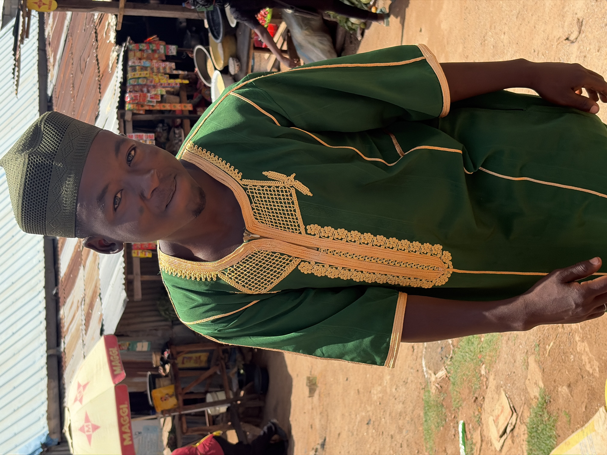 A medium shot depicts a man in a green traditional garment with gold trim and a matching kufi. He is positioned in what appears to be an outdoor market or commercial area. In the background, there's a corrugated metal structure with various items displayed for sale. A "MAGGI" brand sign is visible, suggesting the location is a food stall or shop. The ground appears to be dirt or sand, consistent with a market environment. The image is taken in Barakin Lerawi, Nigeria. The man's pose is relaxed as he looks into the camera. Other people are visible in the background.