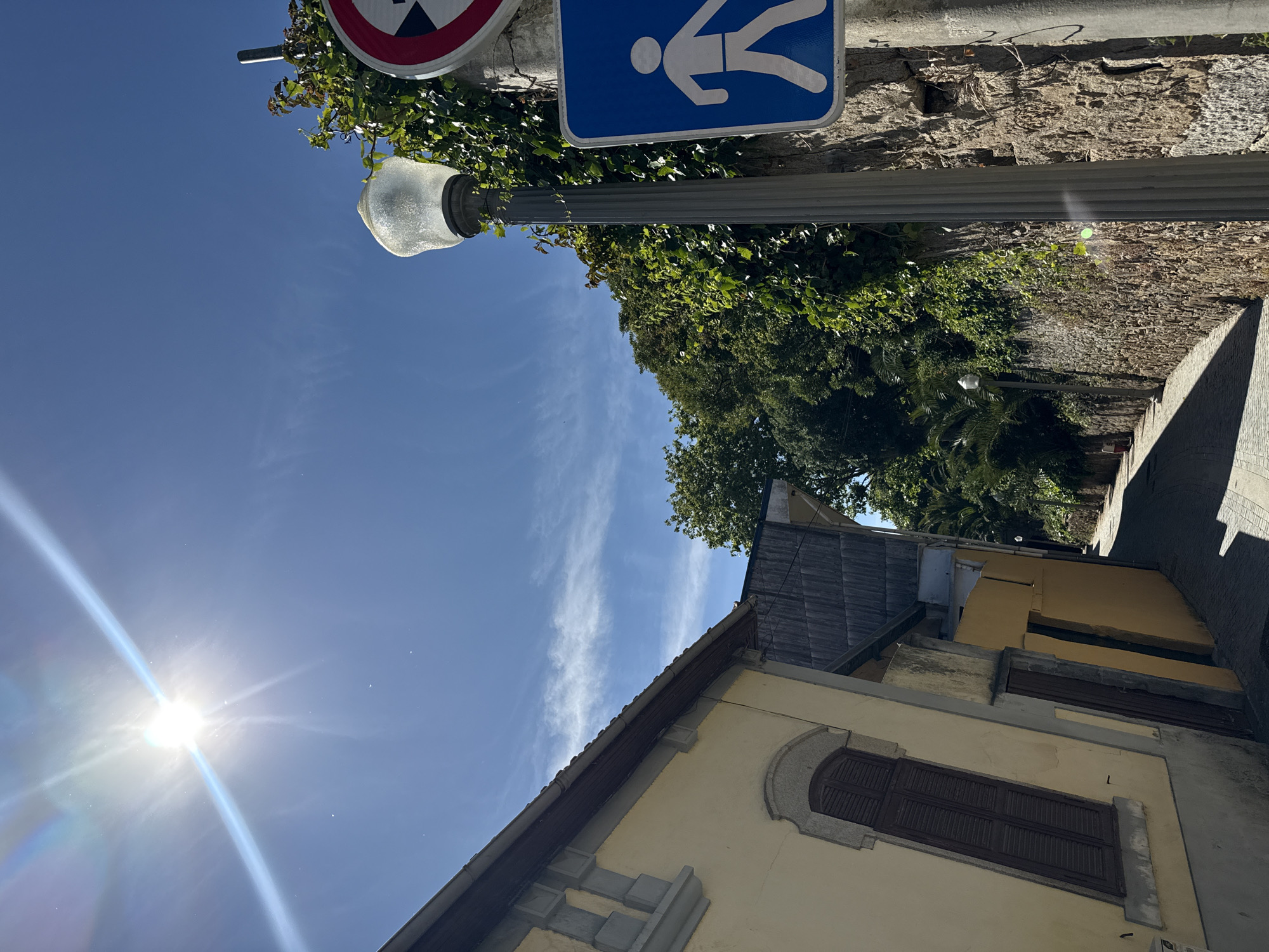 The image presents a low-angle view of a scene dominated by architectural elements and street signs. A portion of a yellow building with a brown shuttered window and gray decorative elements is visible. Above the building, foliage from trees and bushes provides a green backdrop. To the left, a bright sun is visible in the blue sky. A street light, partially obscured by greenery, is attached to a stone wall, which also supports two street signs: one circular with a red and white design, and another rectangular blue sign featuring a white pedestrian symbol. The location of the photograph is Porto, Portugal.
