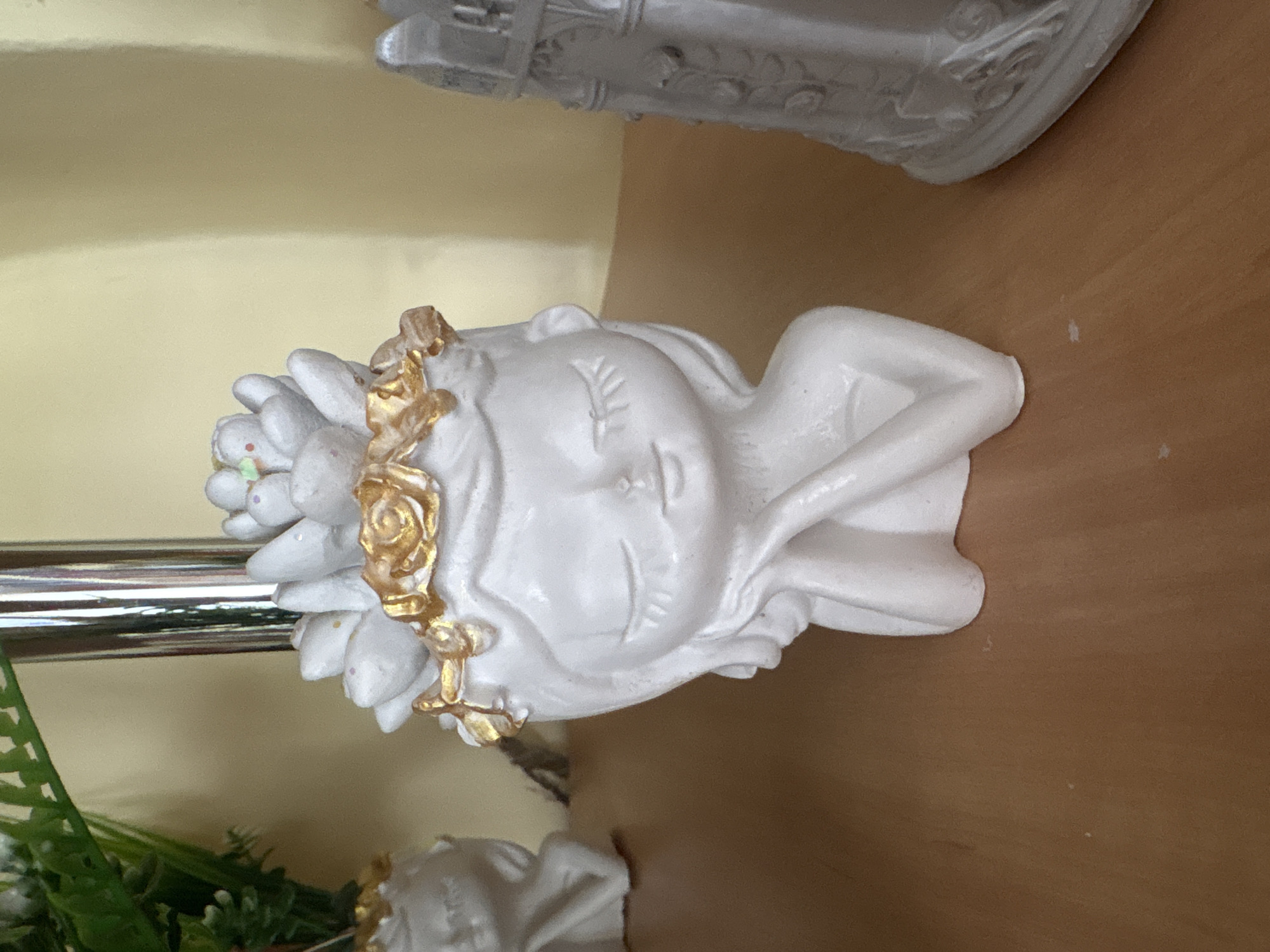 The image shows a white ceramic planter in the shape of a woman's head with eyes closed and a serene expression. She has a delicate golden wreath adorned with floral elements around her head, and her hair appears to be composed of succulent-like shapes. Her arms are resting on her cheeks, giving her a contemplative pose. The planter is placed on a wooden surface, and in the background, there is a partially visible plant in another planter and a silver metallic pole. To the right, another white decorative object, possibly another planter or sculpture, is visible. The lighting suggests an indoor setting with a soft, diffused light source. This scene is located in Ветрынскі сельскі Савет, Belarus.