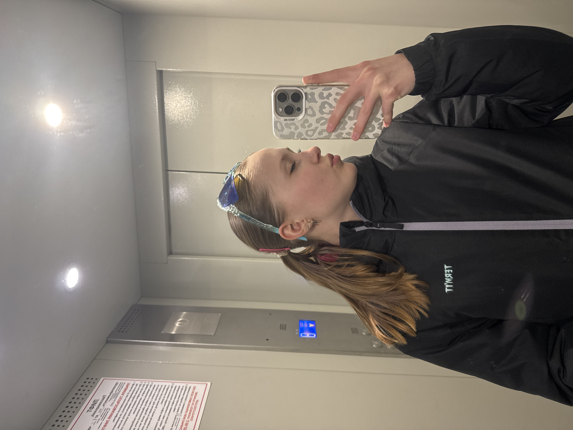 A young woman takes a selfie in an elevator. She holds a smartphone with a gray and white patterned case. She wears a black jacket with a white zipper and a logo that reads "TIMYAET" (likely backwards). Her hair is pulled back into a ponytail, and she wears blue sunglasses on her head. The elevator is metal, with a panel of buttons and a sign in Russian. This photo was taken in the city of Chaikovsky, Russia.