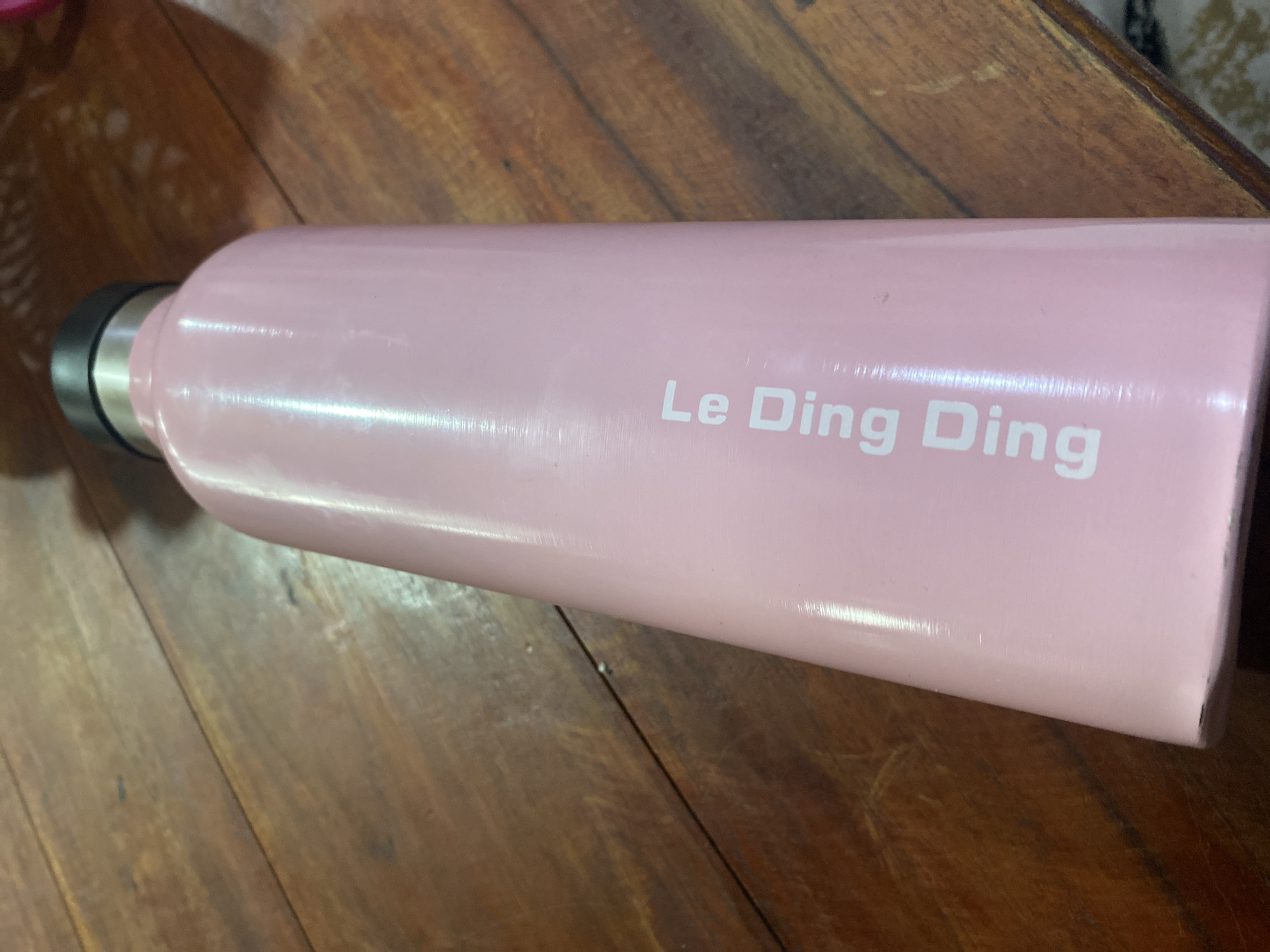 A light pink water bottle with "Le Ding Ding" printed in white text vertically on its side rests on a wooden surface. The bottle has a black cap with a silver band underneath. The wooden surface appears to be a table with visible wood grain and some minor imperfections. The lighting in the image suggests an indoor setting. No specific location in Ta', Indonesia, can be determined from the image.