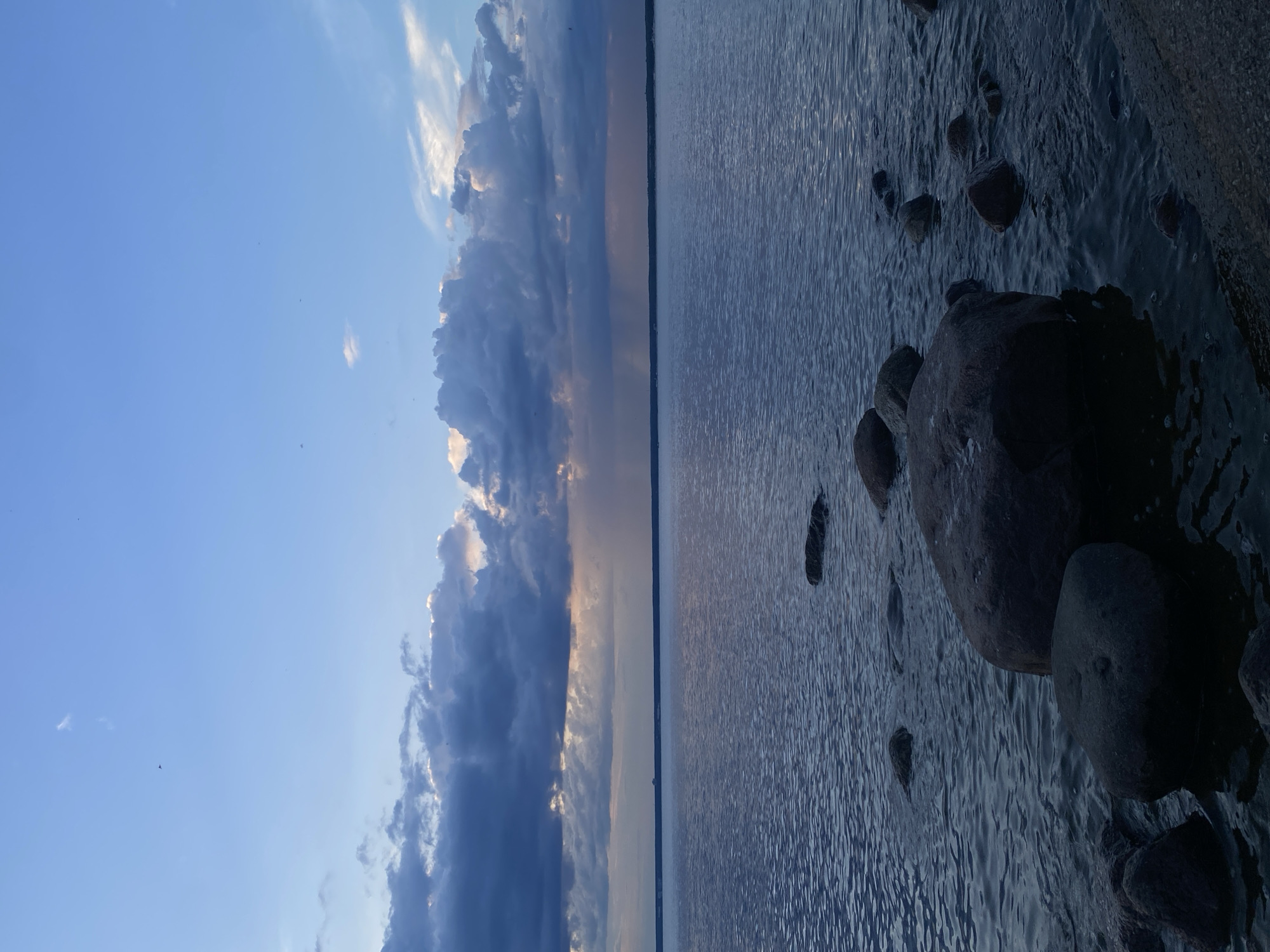 A wide shot captures a body of water in Sestroretsk, Russia, under a cloudy sky with shafts of light breaking through. The water is calm with ripples, and several large, dark rocks are visible along the shoreline, some partially submerged. The sky is a gradient of blue, transitioning to darker, dramatic clouds on the left side of the frame, with a distant horizon line. The overall scene conveys a serene, natural environment.