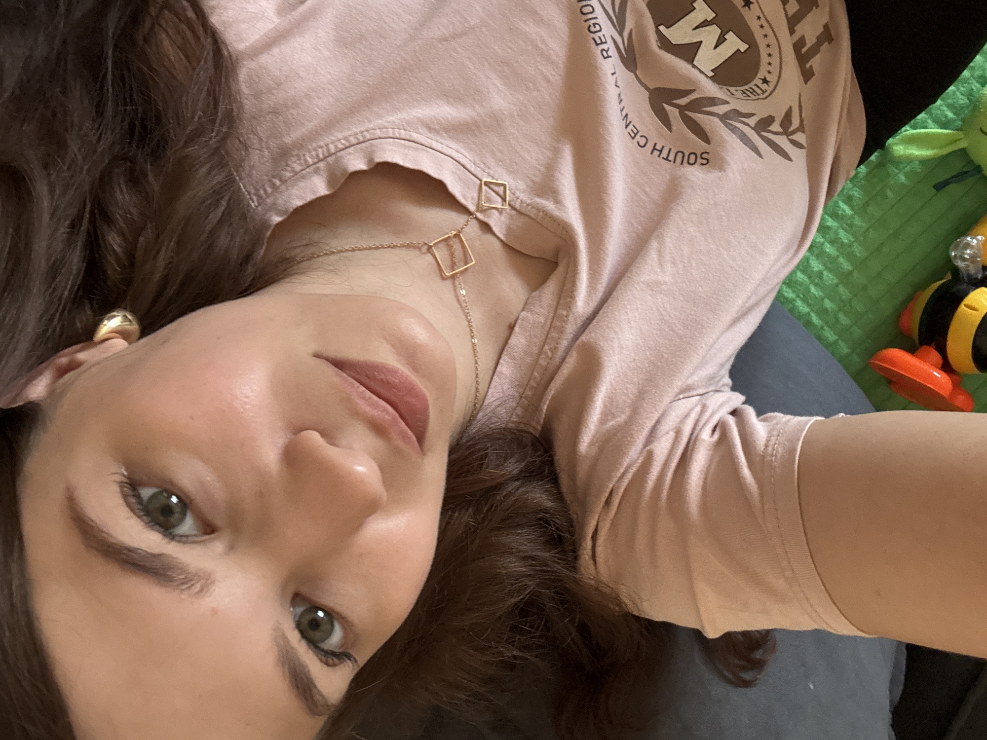 This media file is a close-up selfie of a young woman lying down. She has long, dark brown hair, light skin, and green eyes. She is wearing a pale pink t-shirt with a graphic design on the left side of her chest, which includes a stylized "M" inside a circular emblem and some text. The text visible on the shirt reads "THE SOUTH CENTRAL REGION". She is also wearing a delicate gold necklace with two small square pendants. A gold hoop earring is visible on her left ear. The background is slightly out of focus but appears to be indoors, with a green textured surface and a small, colorful toy that resembles a bee visible on the right side. The lighting suggests it is daytime, possibly indoors with artificial light. The woman's expression is neutral, and she seems relaxed.