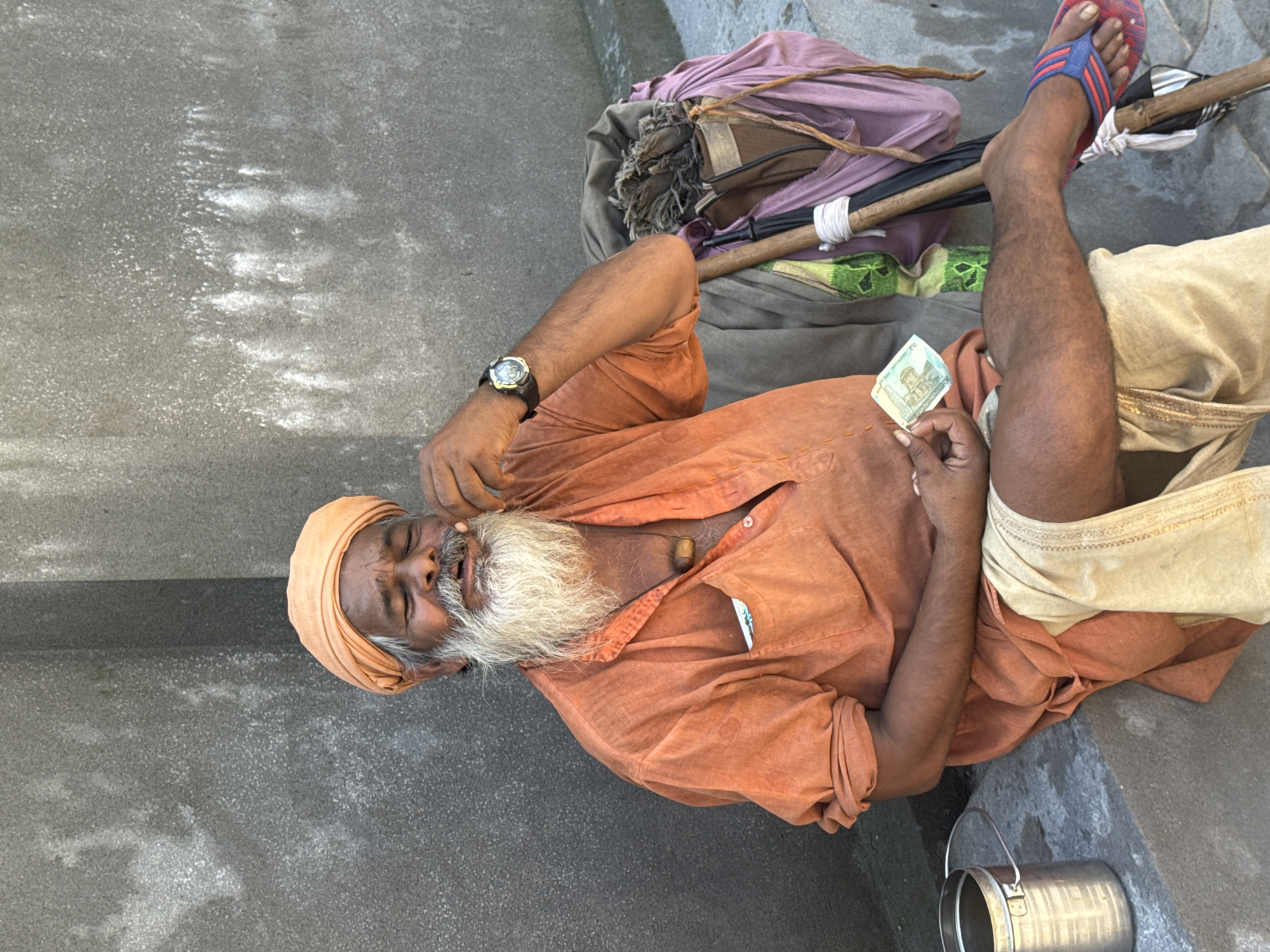 A man wearing a saffron turban and an orange kurta sits on a stone ledge against a gray concrete wall. He has a long white beard and is holding a piece of something to his mouth, with his eyes closed. In his right hand, he holds a banknote. Beside him is a pile of belongings including a wooden staff and a pink bundle wrapped in cloth. To his left is a metal container. The scene appears to be outdoors in a public space, possibly a street or walkway in the city of Rishikesh, India, given the attire and cultural context.