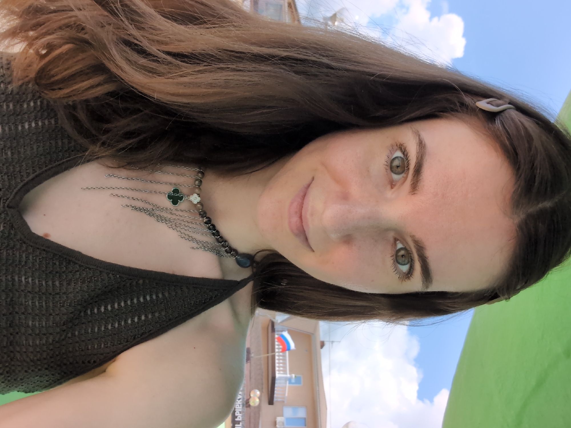 A woman with long brown hair and hazel eyes smiles at the camera outdoors. She is wearing a dark brown crocheted halter top and a necklace with a clover pendant. Behind her, a building with a white balcony and a Russian flag is visible under a cloudy blue sky. The scene appears to be in Divnomorskoye, Russia.