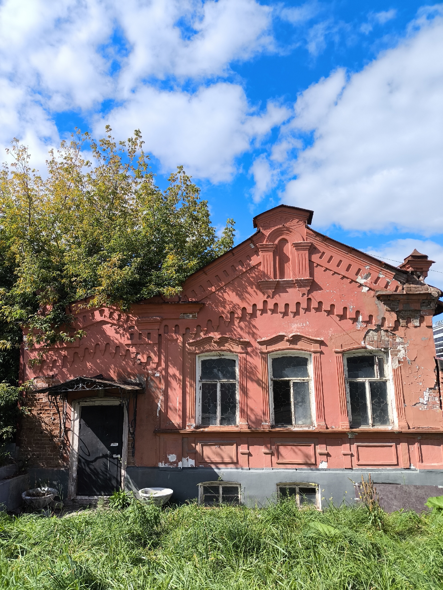 A weathered, salmon-pink, two-story building, likely a former residential house, stands under a bright blue sky dotted with fluffy white clouds. The building features ornate architectural details, including decorative brickwork along the roofline and elaborate window surrounds. Several windows are visible, some with broken panes or boarded up. The façade shows signs of significant decay, with peeling paint and exposed brickwork, particularly on the right side of the building.
A large, leafy tree with yellowing foliage partially obscures the left side of the building, reaching over the roof. The entrance to the building is a dark-colored door under a decorative metal awning. In front of the building, tall, green grass and weeds grow abundantly, reaching waist-high in some areas. Two circular concrete planters, one containing dry leaves, are visible in the overgrown grass near the entrance. The lower level of the building has a gray-painted foundation, and smaller windows are visible at this level.
The image is taken from a low angle, emphasizing the imposing nature of the building and the overgrown foreground. The overall impression is one of neglect and abandonment, juxtaposed against the vibrant blue sky and green foliage. The building appears to be located in Yekaterinburg, Russia, likely in a historical or older district.
