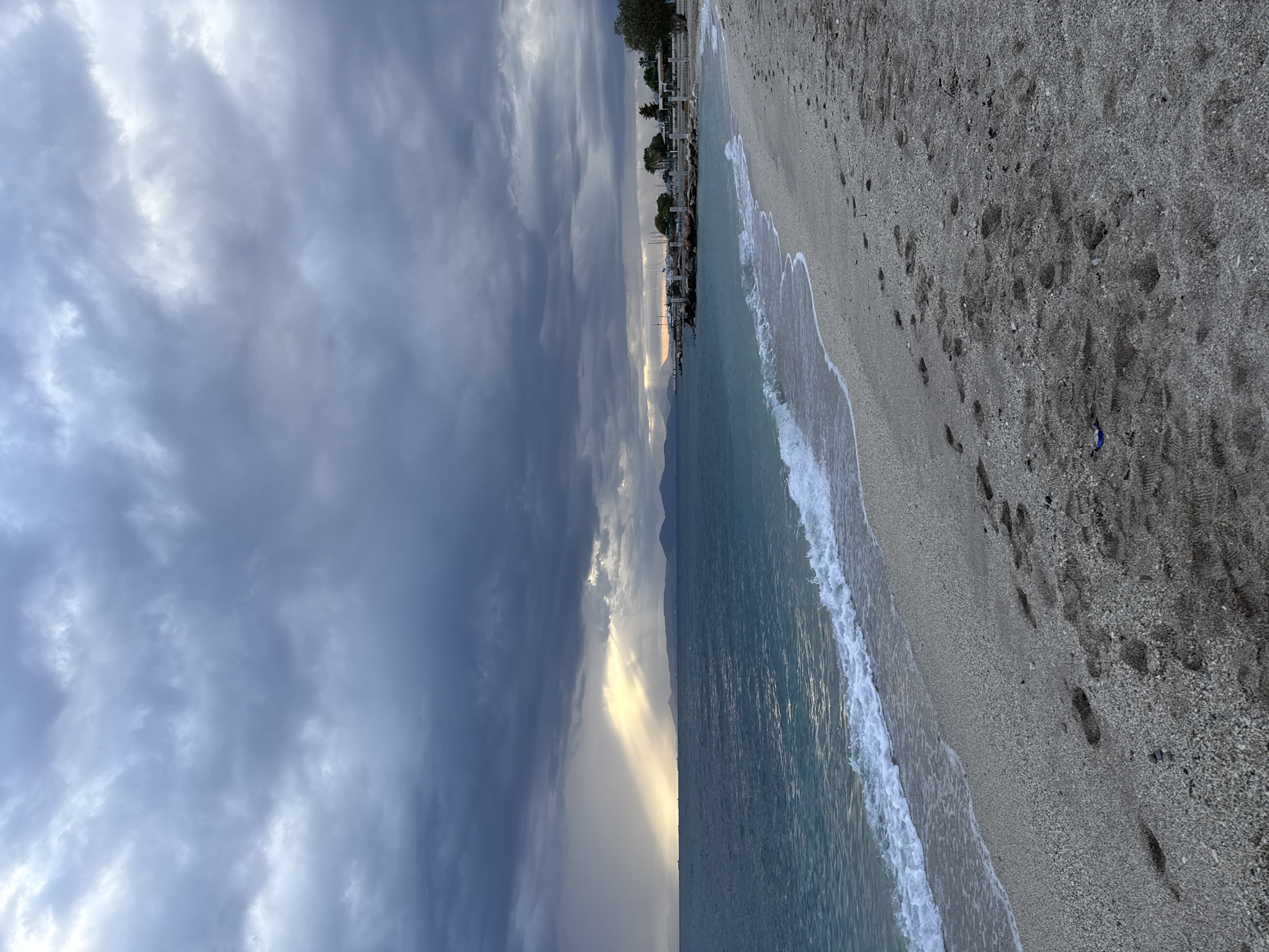 The image depicts a beach scene in Alimos, Greece. The sky is predominantly overcast with heavy, dark clouds. There is a narrow strip of bright light breaking through the clouds near the horizon. The sea is calm with small waves lapping onto the pebble beach. Footprints are visible in the sand. Along the horizon, there appears to be a line of trees and buildings, possibly indicating a marina or harbor area. There are no people visible in the image. A blue piece of litter is visible on the pebble beach.