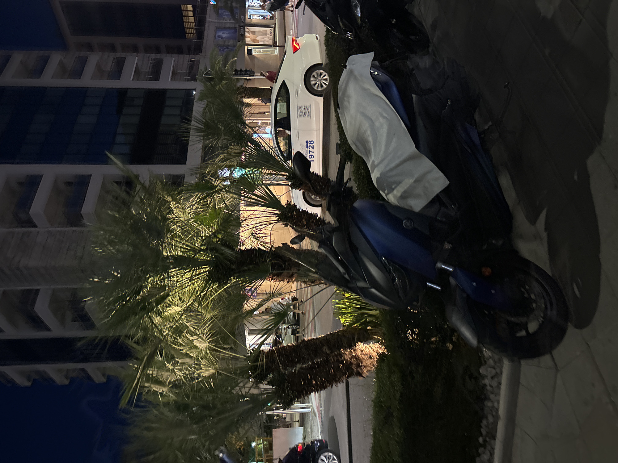 An evening scene in Budva, Montenegro, shows a dark blue scooter parked on a sidewalk in front of lush palm trees. A white sedan, identified as a taxi with the number "19728" visible on its side, is driving down the street. Buildings with illuminated windows and balconies line the background, suggesting an urban setting. The sky is dark, indicating it is nighttime. The street appears wet, possibly from recent rain. People are visible walking on the sidewalk and near the taxi.