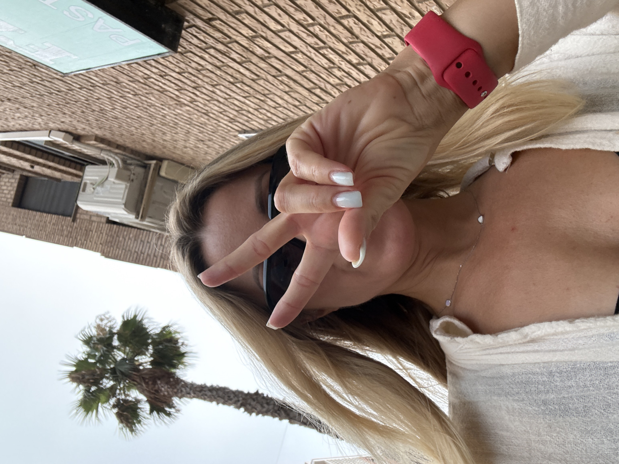 A woman with blonde hair, wearing sunglasses and a white cardigan, is taking a selfie. She is holding up two fingers in a peace sign. Behind her, a palm tree is visible against a cloudy sky. To her right, a brick building with an air conditioning unit and a sign that reads "PAST" can be seen. The scene appears to be outdoors in a city, potentially Valencia, Spain, given the presence of a palm tree and the architectural style. The woman is wearing a red smartwatch on her left wrist and a delicate necklace with a small pendant.