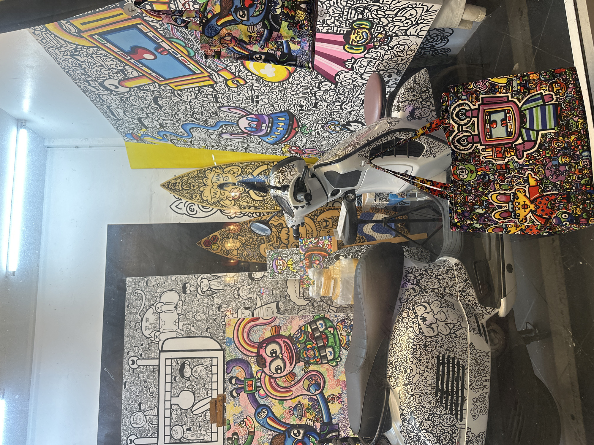 A scooter is parked inside a shop or gallery in Baan Lamai, Thailand. The walls and floor are covered in black and white doodle art with occasional colorful cartoon characters. The scooter itself is also covered in the doodle art style. A colorful tote bag with similar doodle art is placed near the scooter. The scene is illuminated by bright fluorescent lighting from above.
