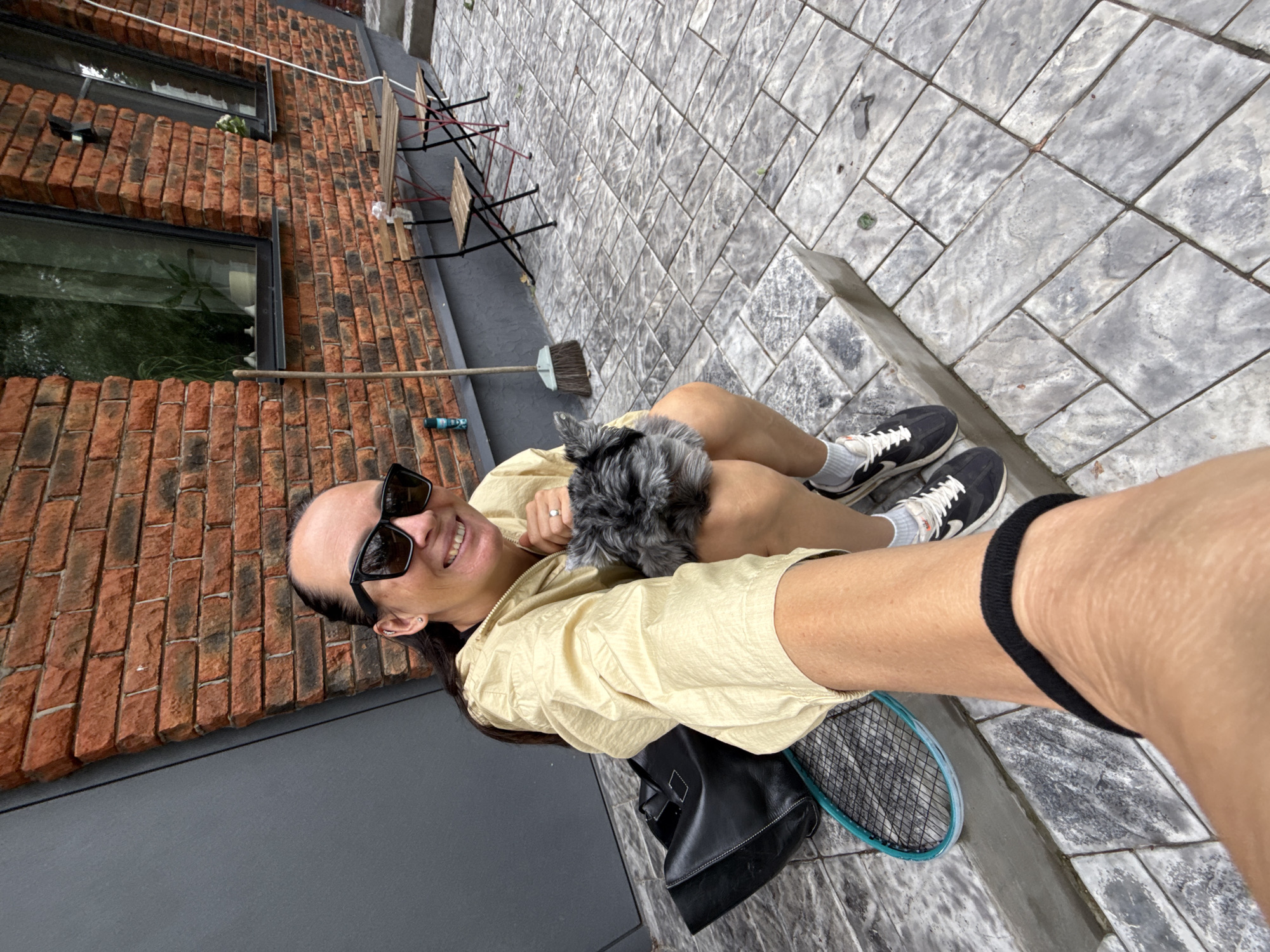 A woman with dark hair pulled back, wearing sunglasses and a light yellow jacket, is sitting on concrete steps outside a brick building, holding a small, grey, fluffy dog in her lap. A black handbag and a tennis racket are visible on the steps beside her. The ground is paved with rectangular grey stones. In the background, a wooden table and chairs are set up near windows in the brick wall. A broom leans against the wall. The scene is outdoors.
