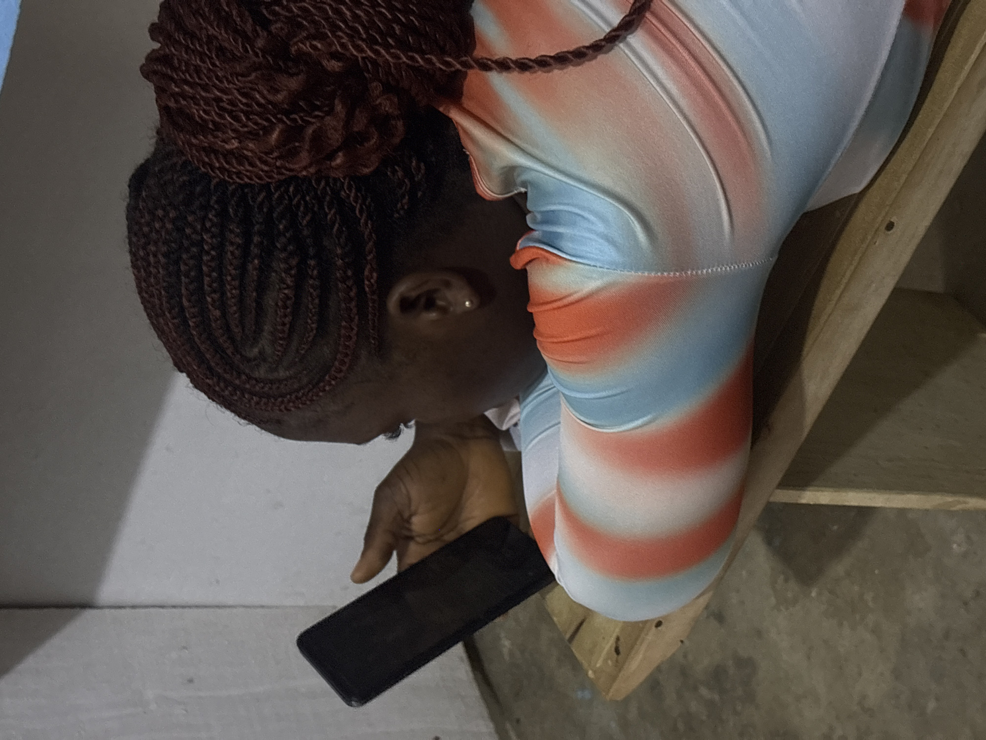 A woman is sitting, seemingly on a wooden structure, and using a black smartphone. Her hair is styled in dark reddish-brown braids, with some gathered in a bun. She's wearing a light blue and white top with orange stripes. The setting appears to be indoors. No details are available to confirm if the location is indeed Ikot Ekpene, Nigeria.
