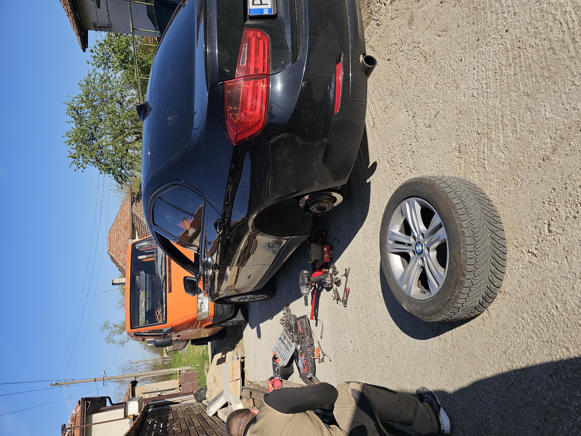 The image depicts an outdoor scene in Krasnovo, Bulgaria, on a bright, sunny day. A black BMW sedan is the central subject, elevated on a red hydraulic jack, with its rear passenger-side wheel removed. The corresponding wheel, featuring a silver BMW alloy rim and an all-season tire, rests on the concrete ground to the right of the car.
A person is observed from behind, crouching near the car and tools, indicating they are in the process of changing or repairing the tire. They are dressed in a beige jacket and dark pants. Numerous automotive tools are scattered on the ground, including a red and black cordless impact wrench, a comprehensive socket set in an open case, and various wrenches and small parts.
In the background, an orange van is parked close to the BMW. Further back, residential buildings with traditional terracotta roofs, green trees, and overhead power lines are visible against a clear, vibrant blue sky. The ground is a light-colored, textured concrete surface.
Notable details include the clear, sunny weather, indicating daytime. A partial view of the BMW's rear license plate shows "BG" (indicating Bulgaria) and parts of a number sequence "E D 4209". The detailed tread pattern of the all-season tire is clearly visible on the detached wheel. The overall setting suggests an informal vehicle maintenance activity taking place in a village or residential area.