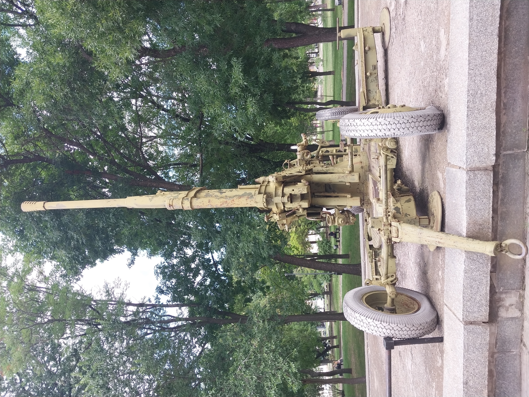 This image displays a military anti-aircraft gun displayed as a monument in an outdoor setting. The gun is painted a muted, olive-drab green and appears weathered, suggesting it is an historical artifact. It is positioned on a concrete and stone base, likely within a park or memorial ground, given the presence of trees and manicured grass in the background. Two large, spoked wheels are visible, supporting the gun's carriage. The scene is set during daylight, with ample natural light illuminating the area. The sky is bright, suggesting fair weather.
The gun itself is the primary object of focus, a long barrel extending horizontally from the main body. Intricate mechanical components, levers, and adjustments are visible, indicating its function as a piece of artillery. The setting suggests a public space, possibly a memorial park in Dnipro, Ukraine, given the provided location context. There is no explicit text visible in the image. The overall mood conveyed is one of historical remembrance and perhaps a somber reflection on military history. The environment appears calm and quiet, with no active events taking place other than the display of the artifact itself.