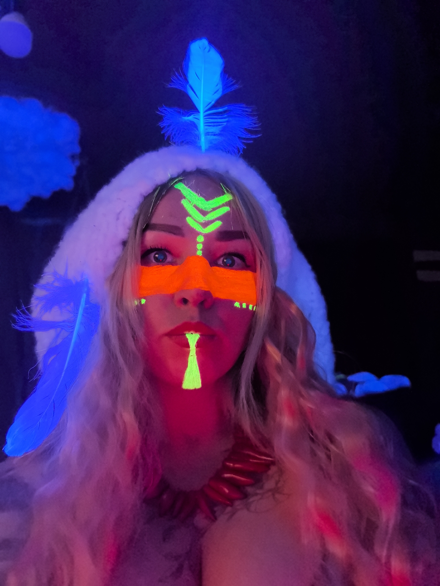 A woman is captured in a close-up portrait, illuminated by UV or blacklight, creating a vibrant and otherworldly aesthetic. Her face is adorned with elaborate glow-in-the-dark paint, featuring bright green chevron patterns on her forehead, a line of green dots down her nose, a bold orange band across her eyes and upper cheeks, and a distinct green vertical marking on her chin. Her eyes are wide and focused directly at the viewer.
She wears a white, fluffy hood, from which several bright blue glowing feathers emanate, with additional blurry blue glowing feathers and a cloud-like shape visible in the dark background. Her long, light-colored hair flows around her, appearing with hints of red and pink due to the intense lighting. Around her neck, she wears a chunky, red, claw-like or beaded necklace. The scene is set in a dark environment, likely an indoor space or night event, emphasizing the luminescence of the neon elements. The image provides no specific internal cues for the given location context of Noviy, Russia, nor does it contain any visible text.