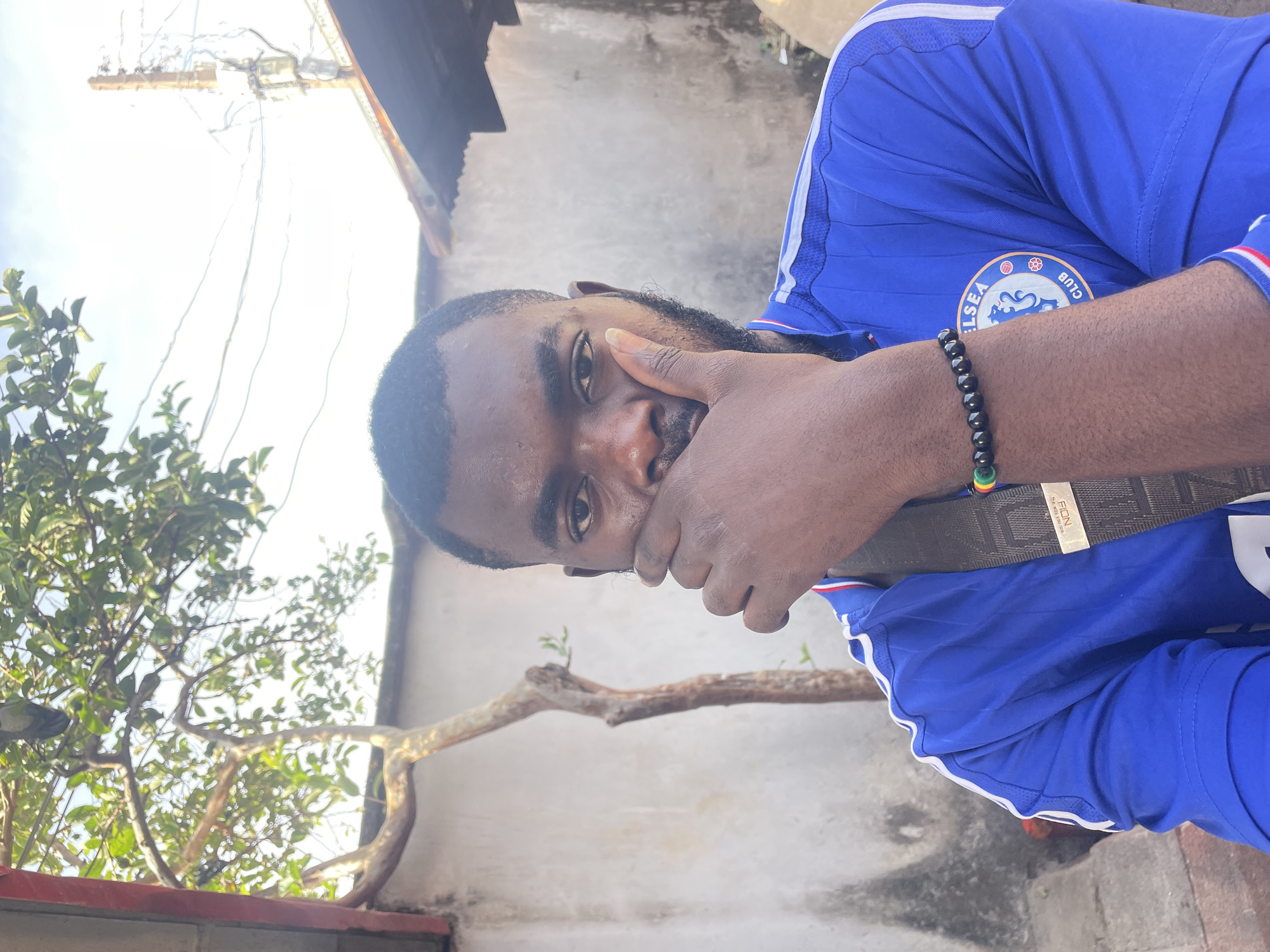 A man with dark skin, wearing a Chelsea soccer jersey and a beaded bracelet, poses outdoors. He holds his right hand to his face, partially covering his mouth. A brown belt is slung across his chest. The environment includes trees, a wall, and overhead electrical wires. The location is plausibly in the city of Jos, Nigeria, based on the style of building and local vegetation.