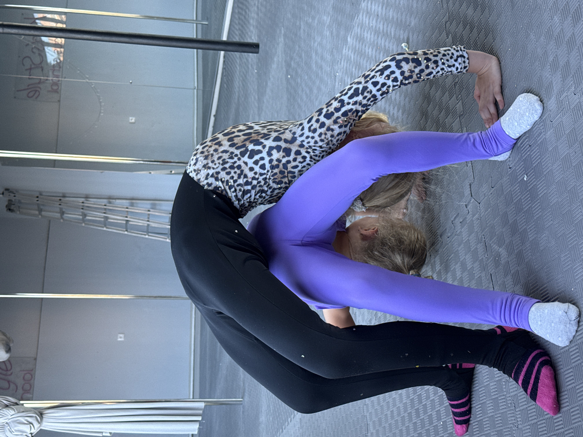 The image shows a young woman performing an extreme contortionist pose on a grey, textured floor. She is bent backwards at a severe angle, with her body forming a U-shape. She is wearing a leopard-print long-sleeved top with rhinestones and black leggings. Her left leg is extended straight, with a grey sock on her foot. Her right leg is bent at the knee, and she is wearing striped socks, one pink and black, and the other black. Her right hand is on the floor, and her left arm is extended upwards, bent at the elbow.
The background appears to be an indoor studio with a wall and some vertical poles. There are reflections on the wall, suggesting it might be a mirrored surface. The lighting is bright, indicating it could be daytime. There is some faint text visible on the wall, which appears to be a logo or name, but it is difficult to read clearly. One visible word fragment appears to be "school". The overall impression is of a skilled athletic performance in a dedicated practice space. The pose suggests strength, flexibility, and dedication.