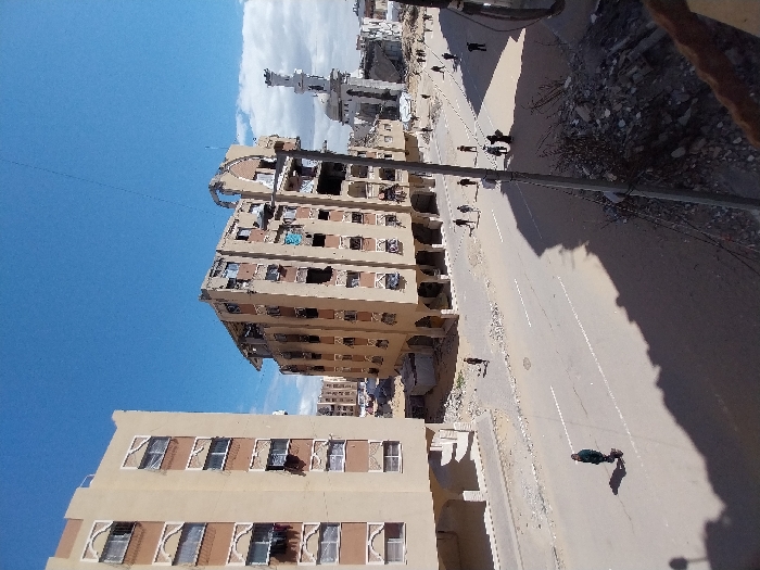 This image depicts a damaged urban street scene in Khan Yunis, Palestinian Territory, under a clear blue sky with scattered clouds. In the foreground, a substantial shadow from an unseen structure falls across the paved road, which features faded white lane markings. To the immediate right, a pile of rubble and debris is present, with wires extending from it.
On the left side of the street, a multi-story, light beige residential building stands mostly intact, showing some open windows and laundry hanging. Across the street, a prominent multi-story, light brown building exhibits severe structural damage, with its upper floors significantly compromised, windows missing, and internal elements exposed. A utility pole or structural column stands directly in front of this damaged building.
Further down the street, on the right side, the remnants of a mosque are visible, characterized by a white dome and a partially damaged, slender minaret. Beyond these structures, more partially destroyed buildings are evident.
Approximately 15-20 individuals are dispersed across the street and sidewalks. One person, dressed in dark clothing, walks alone in the left lane of the road. Other individuals and small groups are seen walking further down the street on both sides, navigating the scene amidst the damaged environment.