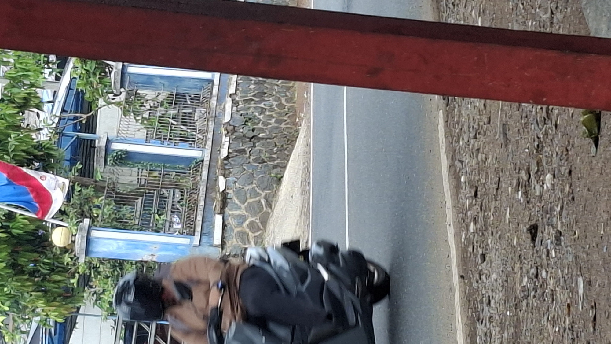 A person wearing a black helmet and a brown jacket is riding a grey scooter on an asphalt road. The scooter is in motion, indicated by a slight blur. To the left of the road, there is a low stone wall topped with a blue and white fence. Behind the fence, there are trees with green leaves and a building with a blue roof. A flag with red, white, and blue stripes is attached to a pole above the fence. To the right of the scene, a red pole is partially visible, obscuring some of the background. The ground on the left side of the road is covered with small stones and dirt.