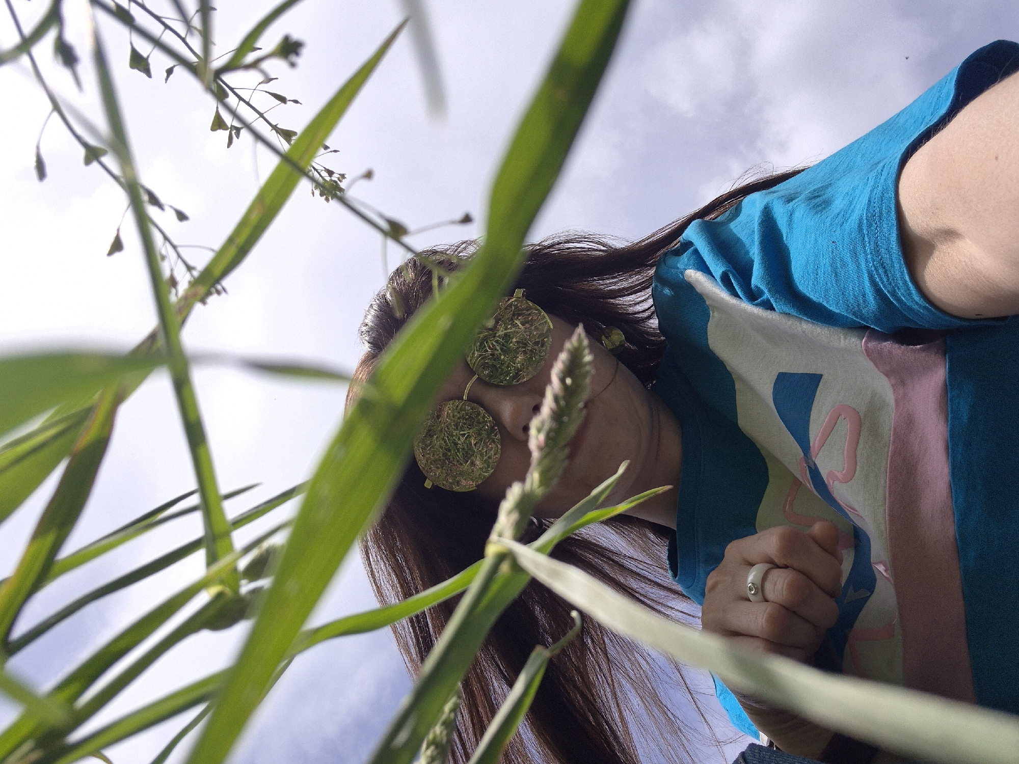 A low-angle shot features a person with long brown hair, wearing round sunglasses filled with what appears to be grass or moss. The person is wearing a turquoise t-shirt with a design that includes pink and white elements. Their right hand, adorned with a white ring, is clenched into a fist and positioned towards the bottom of the frame. Blades of green grass and stalks with small, indistinct seed heads obscure parts of the image, creating a foreground blur. The sky, with scattered clouds, forms the backdrop. The city and specific location are not determinable from the image.
