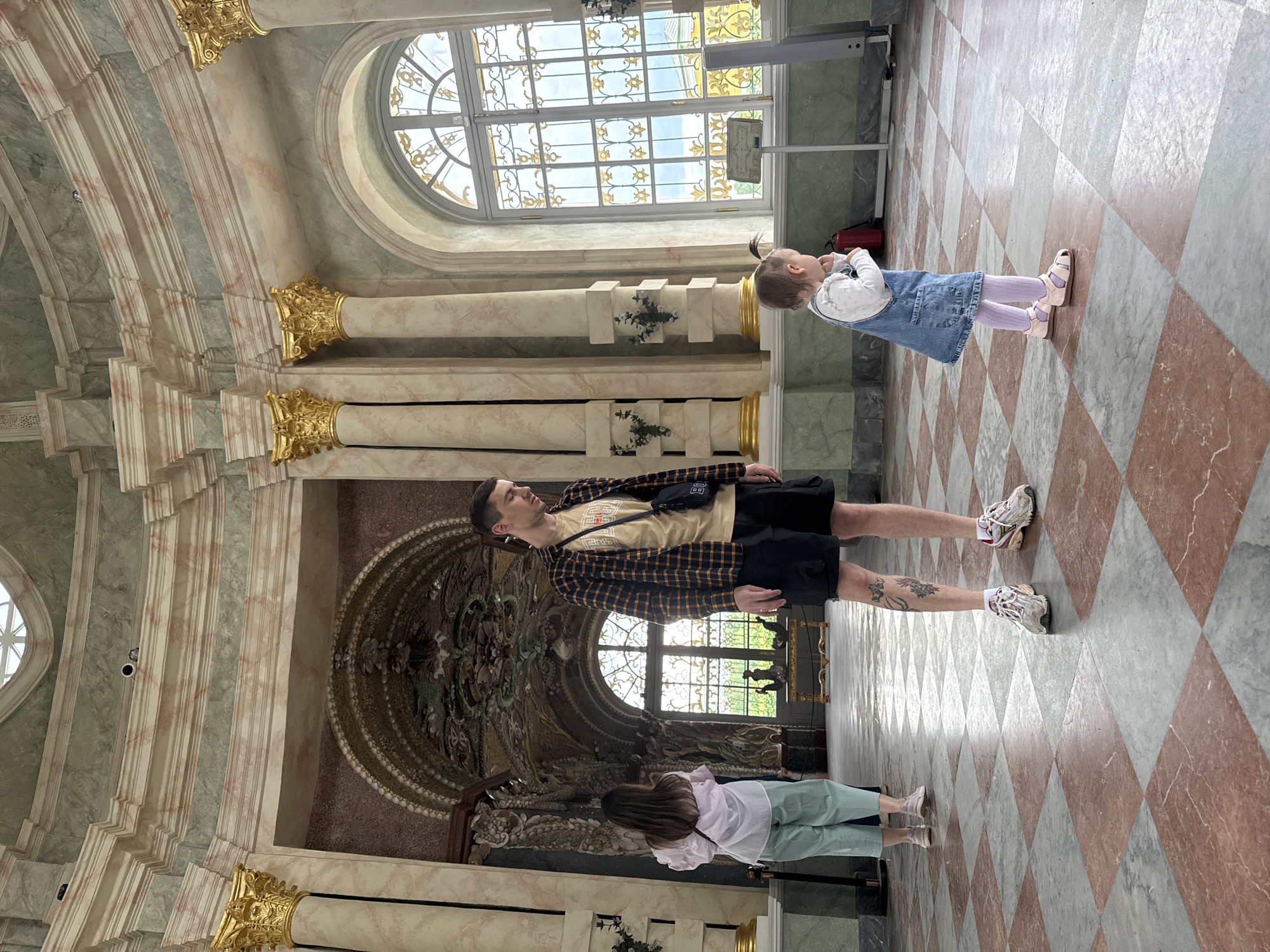 A family is exploring an ornate building interior, potentially a historic palace or museum. The flooring is a checkered pattern of red and grey stone tiles. The walls are finished in pale green marble, adorned with gilded trim. Tall windows punctuate the space, providing natural light. The ceiling is vaulted, exhibiting intricate architectural details.
A man wearing a yellow t-shirt, a black and gold plaid shirt, and black shorts, is standing centrally in the frame. Next to him is a child wearing a denim dress and pink tights. Another woman, wearing a white shirt and mint colored pants, is in the bottom of the frame.
The location is likely within a historical building or palace in Moscow, Russia, given the architectural style and décor.