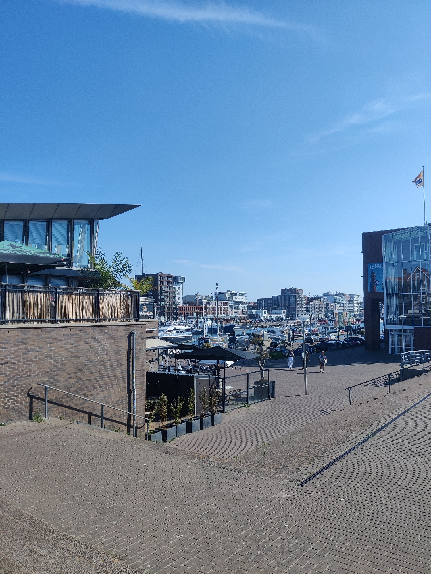 The image depicts a sunny day at a marina in Scheveningen, The Hague, Netherlands. The foreground features a brick-paved walkway and stairs. To the left, a brick building with a modern, glass-fronted upper level and a thatched-style fence runs along the side. Overhanging this building are two large, dark umbrellas, one with a green Heineken logo. Beyond the building, a marina is visible with numerous docked sailboats and other vessels. In the background, a cityscape of apartment buildings and other structures lines the waterfront. To the right, a modern glass-fronted building displays a large blue and white sign that reads "TE HUUR" (For Rent) and other text in Dutch. Several people are walking on the paved area in front of this building, and cars are parked in the vicinity. A flag is visible on a flagpole atop the glass building on the right. The sky is clear and blue with a few wispy clouds.
