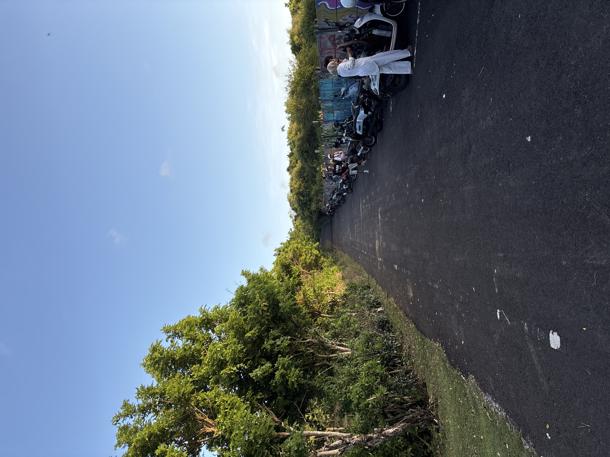 A man dressed in white stands beside a line of parked scooters on a black asphalt road, situated in Kuta Selatan, Indonesia. Vegetation lines the left side of the road, with a blue sky visible above. The scooters are parked near a wall with graffiti. Other individuals can be seen further down the line of scooters.