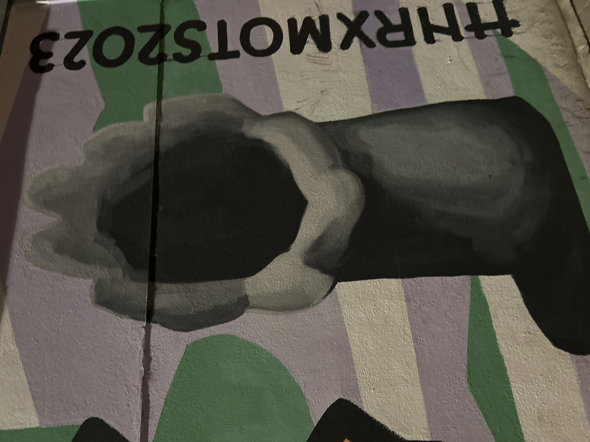 This detailed image captures a section of a mural on a textured wall in Strasbourg, France. The central element is a vertical, abstract form rendered in gradients of black and light grey, featuring a deep, dark recess at its core and contoured edges that suggest a rugged, organic structure. A distinct shadow extends from its base onto the striped background. The wall's background is composed of horizontal bands of light grey, light purple, and green. A partial, dark green, curvilinear shape is visible on the far left. On the right side of the mural, the black, sans-serif text "#HRXMOTS2023" is painted vertically, ascending from bottom to top. The paint exhibits visible brushstrokes and texture, consistent with a painted surface.