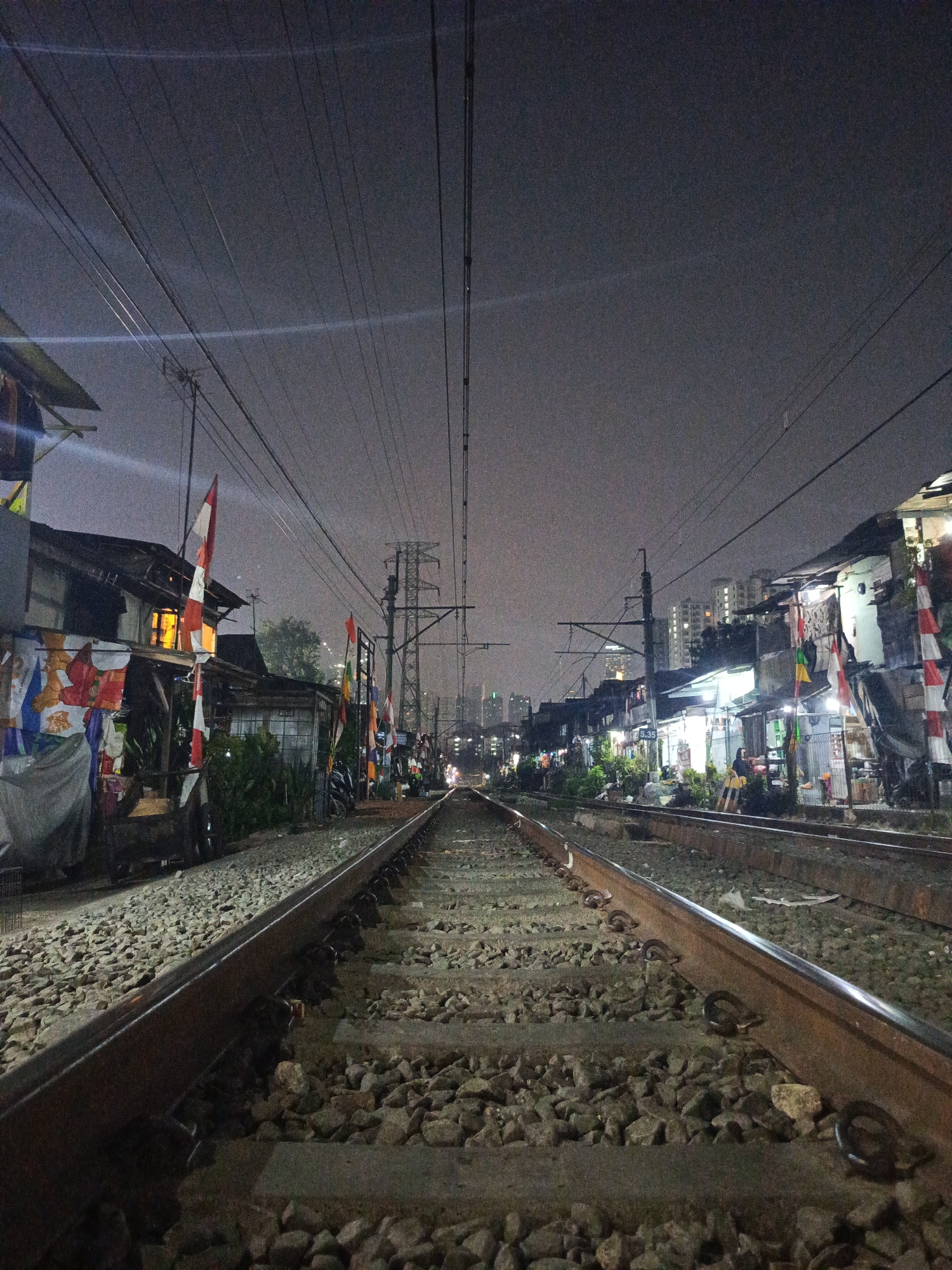 The image depicts a view along a railway track at night in Central Jakarta, Indonesia. The tracks stretch into the distance, flanked by informal settlements on either side. The buildings in these settlements are constructed from a mix of materials, with corrugated metal and makeshift additions visible. Many of the houses are illuminated from within, casting a warm glow onto the scene. Indonesian flags are displayed prominently outside some of the residences. Overhead, a complex network of electrical wires and poles crisscrosses the sky. In the background, the silhouette of a modern city skyline, with taller buildings, is visible against the night sky. Several people are present, with figures seen near the houses and in the distance along the tracks. The ground is covered in ballast stones, typical for a railway environment. The overall atmosphere is one of a densely populated urban area at night, with the railway line serving as a central feature dividing the informal settlements.