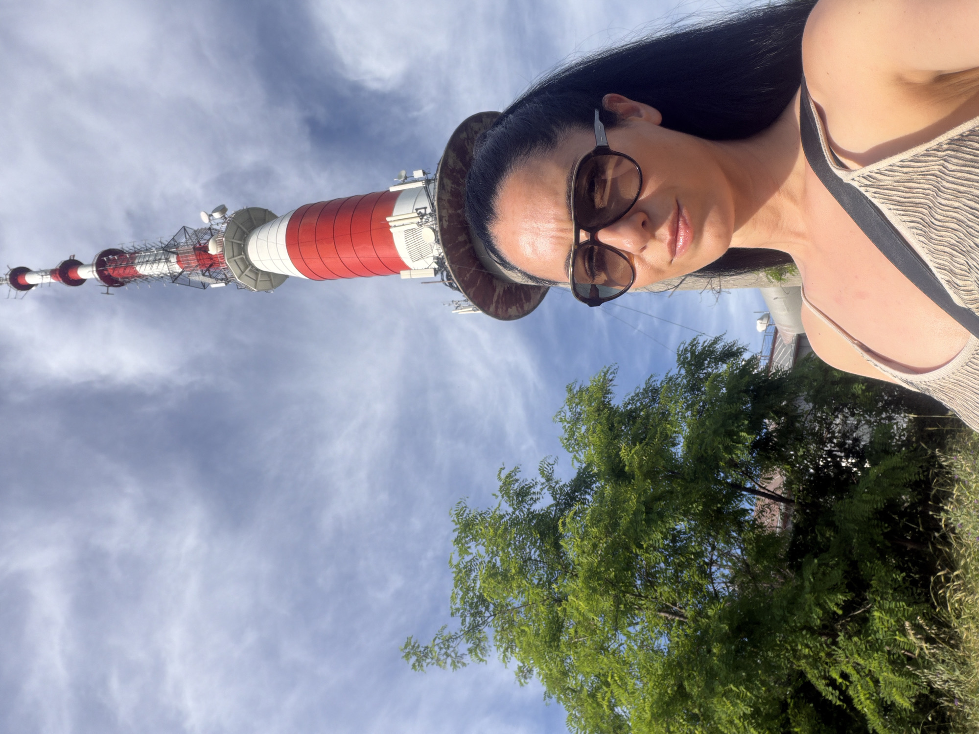A low-angle, medium shot captures a woman with dark hair and sunglasses posing for a selfie in front of a towering red and white broadcast antenna in Pleven, Bulgaria. The woman is wearing a beige, ribbed tank top and has a black strap across her shoulder. The antenna, a prominent landmark in the city, dominates the background, its red and white sections alternating as it stretches towards a sky filled with wispy clouds. To the left of the frame, a lush green tree with dense foliage provides a natural contrast to the industrial structure. The scene is set outdoors on a sunny day, with hints of buildings and vegetation visible in the lower parts of the image.