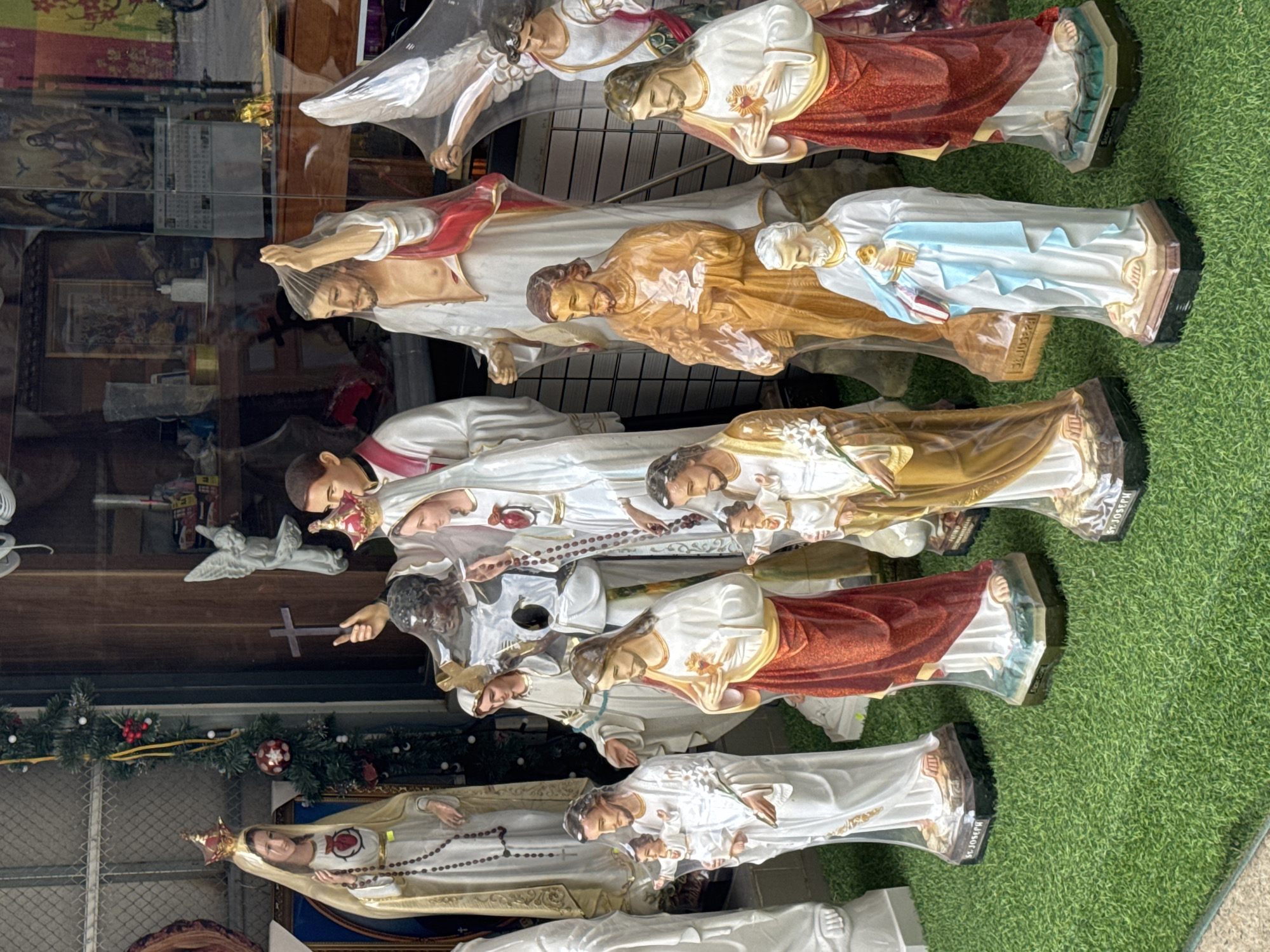 The image depicts a shop window display in Da Lat, Vietnam, featuring multiple religious statues, including figures of Jesus Christ, Mary, and saints. The statues are encased in clear plastic packaging and arranged on what appears to be artificial grass inside the window. Reflections in the glass show elements of the street scene and interior of the shop, including a Christmas garland and religious paintings or prints. The statues are diverse in color and attire, some featuring rosaries and other religious symbols.