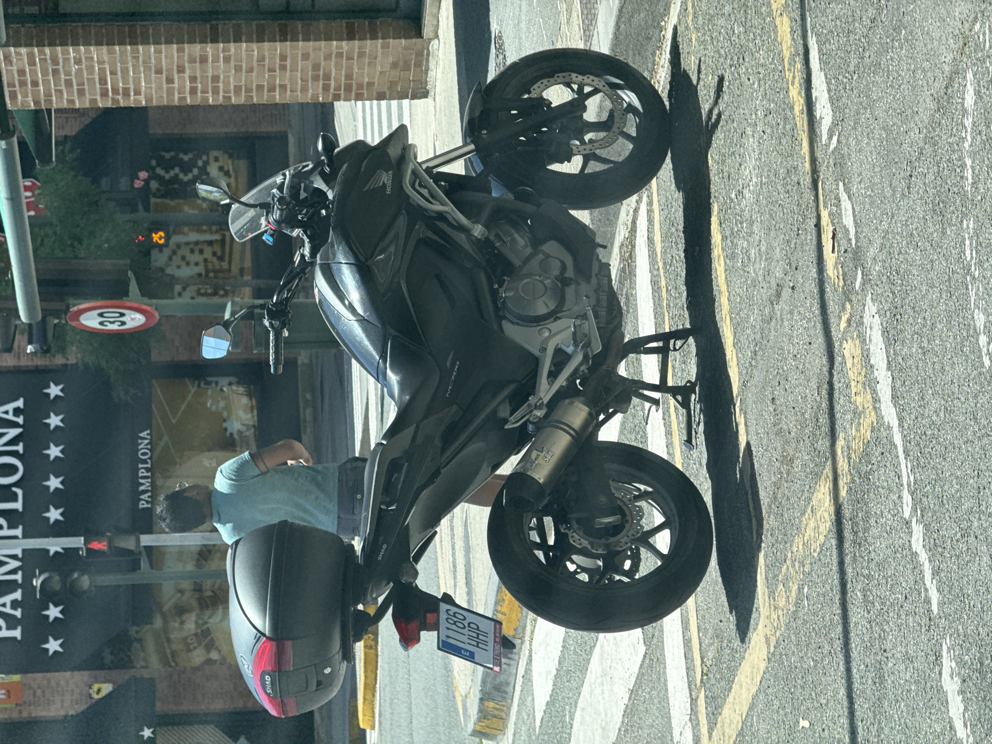 The image shows a parked black Honda motorcycle on a street in Pamplona, Spain. The motorcycle has a black SHAD top box, a license plate that reads "E 1186 HHP", and a silver exhaust pipe. A person in a light blue t-shirt and denim shorts is standing behind the motorcycle, facing away from the camera. In the background, there is a building with a sign that says "PAMPALONA" in large letters, with stars above and below. A speed limit sign indicating "30" is visible on a traffic pole, as well as a pedestrian traffic signal showing a red walking figure. The street is paved, with yellow and white painted lines marking pedestrian crossings and traffic lanes. The scene appears to be a sunny day.