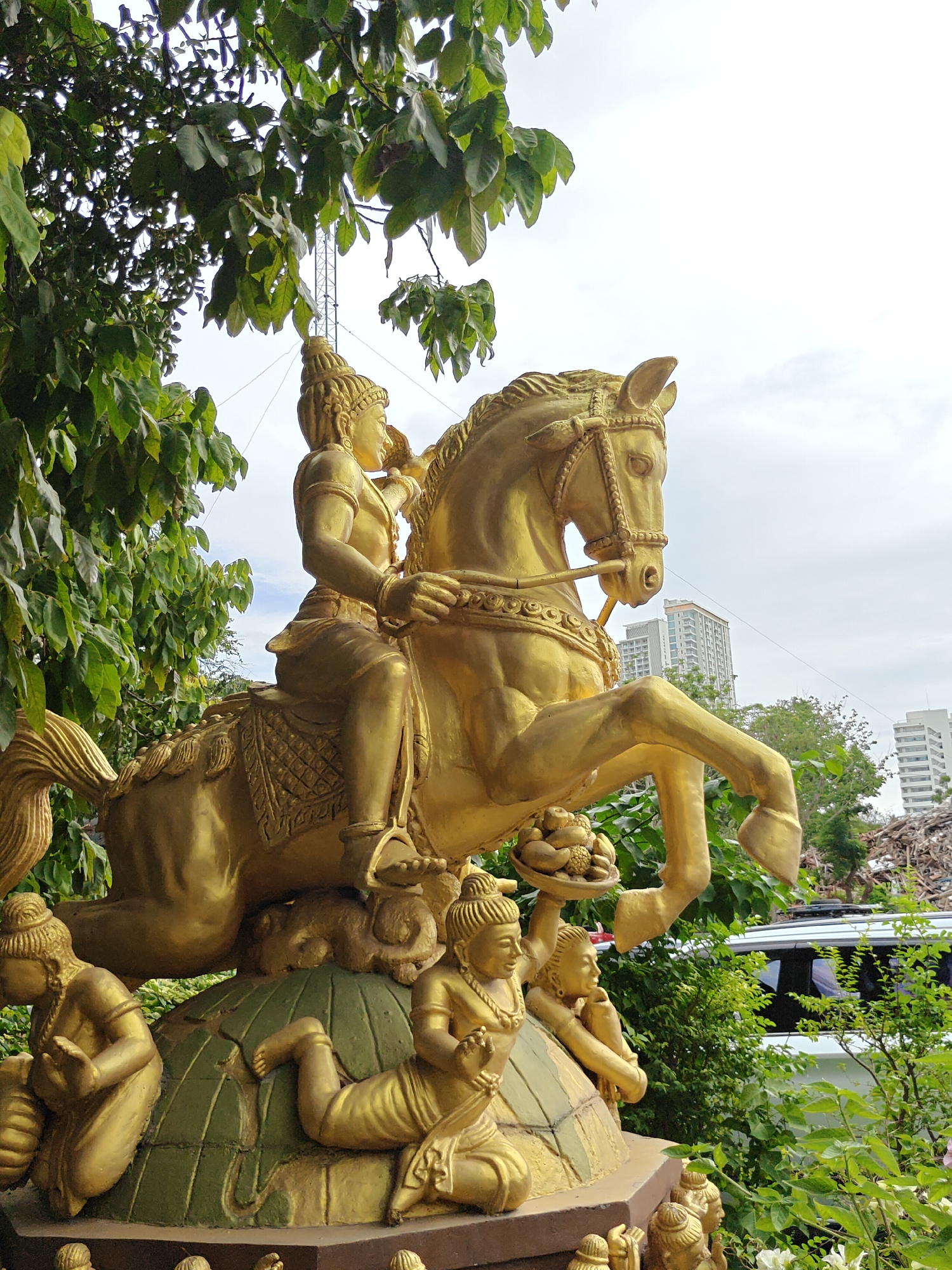 A golden statue of a figure riding a horse is prominently displayed in Pattaya City, Thailand. The statue depicts a person wearing a regal headdress and ornate clothing, seated atop a horse with elaborate tack. The horse is shown in motion, with its front legs raised. At the base of the statue, several smaller golden figures are depicted, some kneeling and others reclining. They appear to be offering or holding items.
The statue is surrounded by lush green foliage, with large leaves obscuring parts of the background. In the distance, modern buildings with multiple stories are visible, suggesting an urban setting. A white vehicle is partially visible behind the foliage, and to the right, a pile of debris or construction materials can be seen. The sky is overcast, indicating a cloudy day. The overall impression is of a cultural or religious monument within an urban landscape.