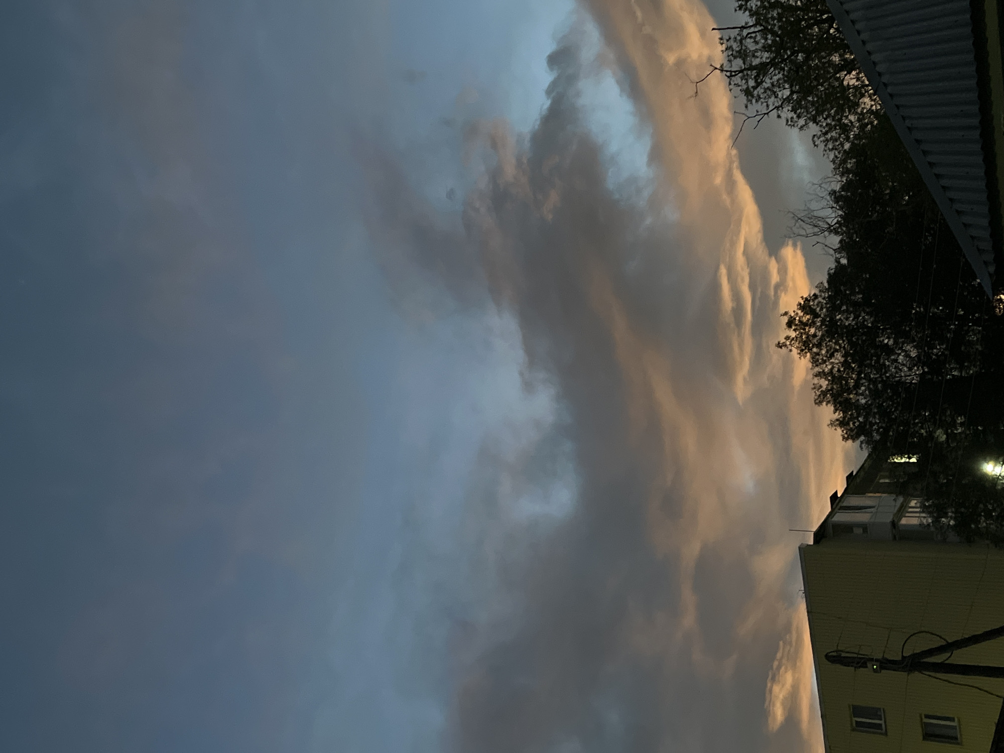 A low-angle, wide shot captures a dusky sky over a residential area in Petropavlovsk-Kamchatsky, Russia. The sky is a blend of deep blue and soft orange, with dramatic, wispy clouds catching the last light of day. In the foreground, a portion of a pale yellow building is visible on the left, featuring several windows and an attached utility pole with wires. Dark silhouettes of trees and a corrugated metal roof partially obscure the view on the right. The scene is devoid of any visible human activity.