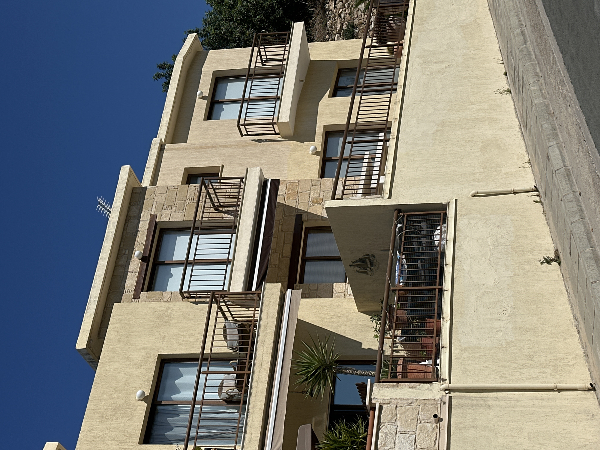 This image shows a section of a multi-story building with a light yellow stucco exterior and stone accents, situated in Peyia, Cyprus. The building has multiple balconies with dark brown railings. On the first balcony visible, there are two chairs with grey cushions. A large window with sheer curtains is behind the chairs. Another balcony above this one has a similar arrangement. Further to the right, another balcony extends out from the building, with a large sliding glass door. The building is positioned on a hillside, with a steep incline leading up to it from a paved sidewalk and road in the foreground. There are potted plants and a small palm tree visible on the balconies and near the building's base. The sky is clear and blue. The overall impression is of a residential or vacation property, common in the coastal areas of Cyprus.