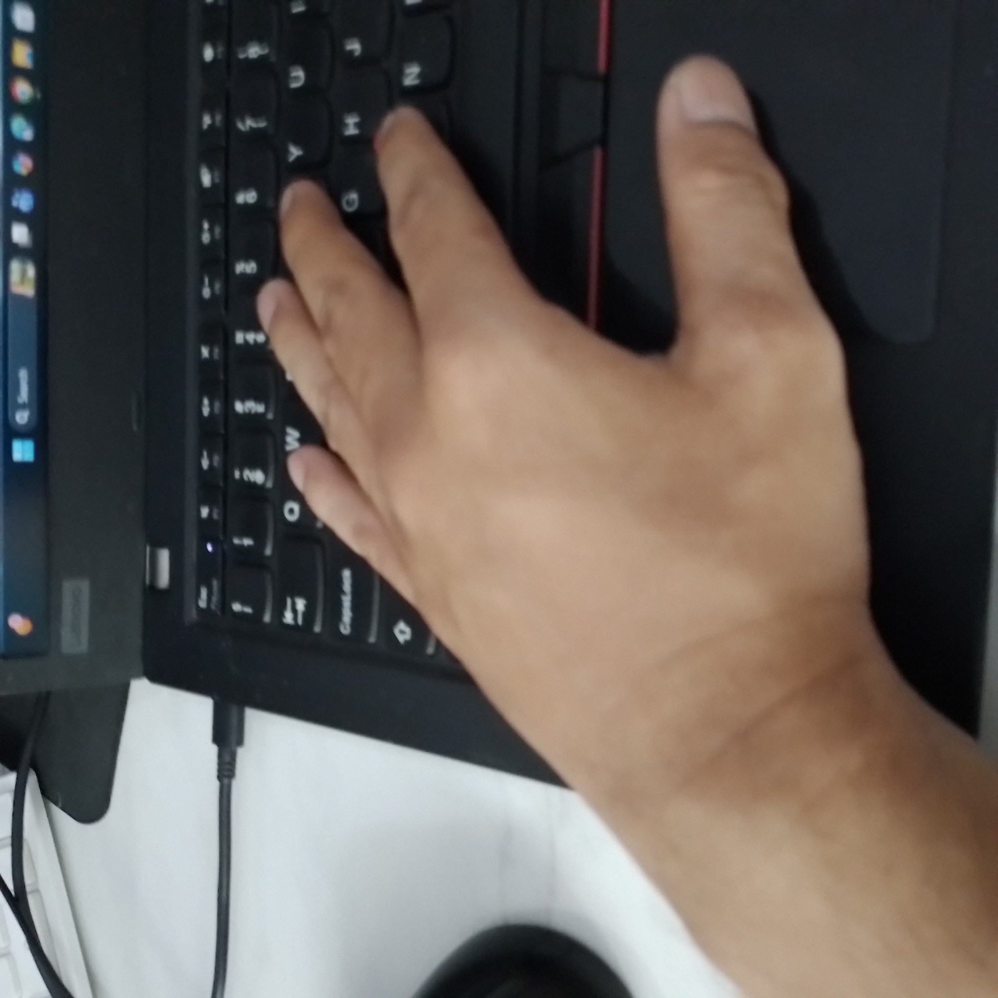 A hand is resting on a laptop keyboard. The laptop is black and appears to be open. The keyboard has a standard QWERTY layout, with some keys visible including "Q", "W", "E", "R", "T", "Y", "U", "I", "O", "P", "A", "S", "D", "F", "G", "H", "J", "K", "L", "Z", "X", "C", "V", "B", "N", "M", and "CapLock". The hand is positioned on the middle and right side of the keyboard, with the index finger resting near the "W" key. The background is blurred and appears to be a white surface, likely a desk. A black cable is plugged into the side of the laptop. The image does not contain any information that would indicate its location in Surakarta, Indonesia.