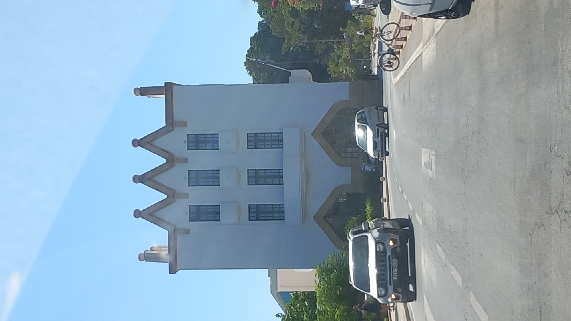 The image shows a street scene in the city of Kos, Greece. A white, multi-story building with a distinct crenellated roofline and arched ground-floor entrances dominates the center of the frame. The building has multiple windows on its upper floors. In front of the building, a silver Jeep Wrangler is driving towards the viewer. To the right of the Jeep, a silver compact car is also driving towards the viewer. The street is paved and has white lane markings. On the right side of the road, a white bicycle is parked next to several orange traffic cones. Trees and foliage are visible behind the building and on the sides of the street. The sky is clear and blue. A person is visible walking on the sidewalk to the left of the Jeep.