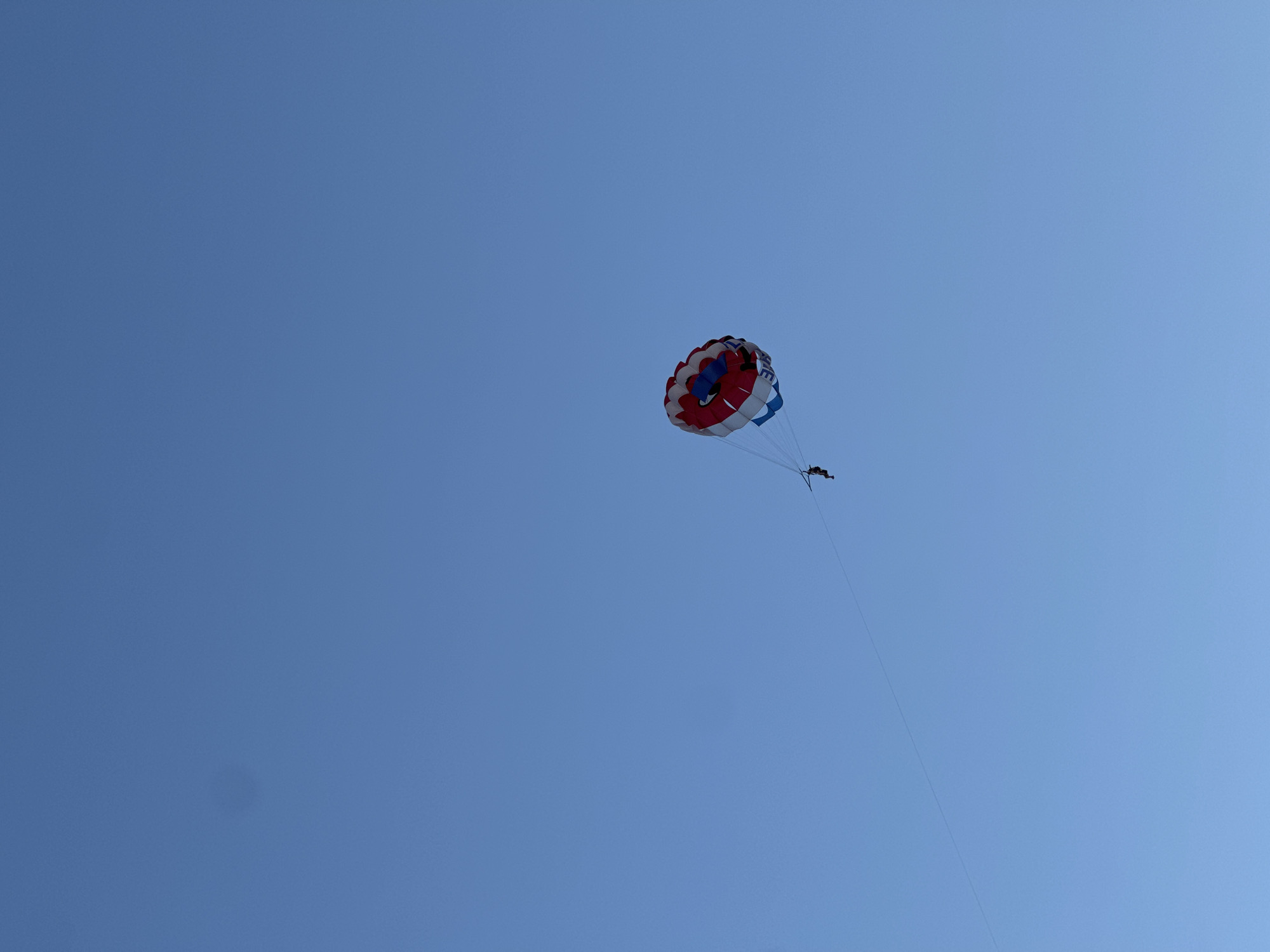 A paraglider with a red, white, and blue parachute is suspended high in a clear blue sky. The parachute is shaped like a dome and has a graphic of a smiling cartoon face on it. A single person is attached to the parachute via a harness and appears to be in a seated position. A thin rope or tow line extends from the parachute downwards and to the left, suggesting it is being pulled by a boat or vehicle. The paraglider is the sole subject in the frame, with the vast expanse of the sky serving as the backdrop. The image provides no direct visual cues to its specific location within Budva, Montenegro, but the activity itself is commonly associated with coastal tourist destinations like those found in Budva.
