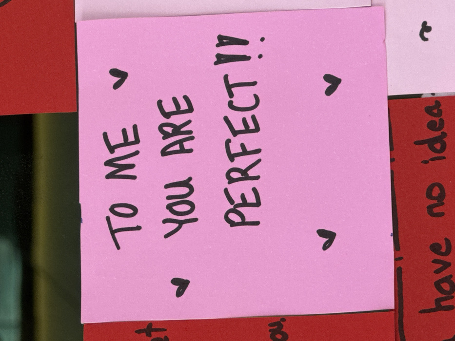 A square, pink sticky note displays a heartfelt message written in black marker. The text reads "TO ME YOU ARE PERFECT!!" and features small heart symbols above and below the words. The note is surrounded by other colorful sticky notes in red and white. A handwritten message "have no idea" is visible on a red note to the right. The image is casually captured, with uneven lighting and a slightly tilted perspective.