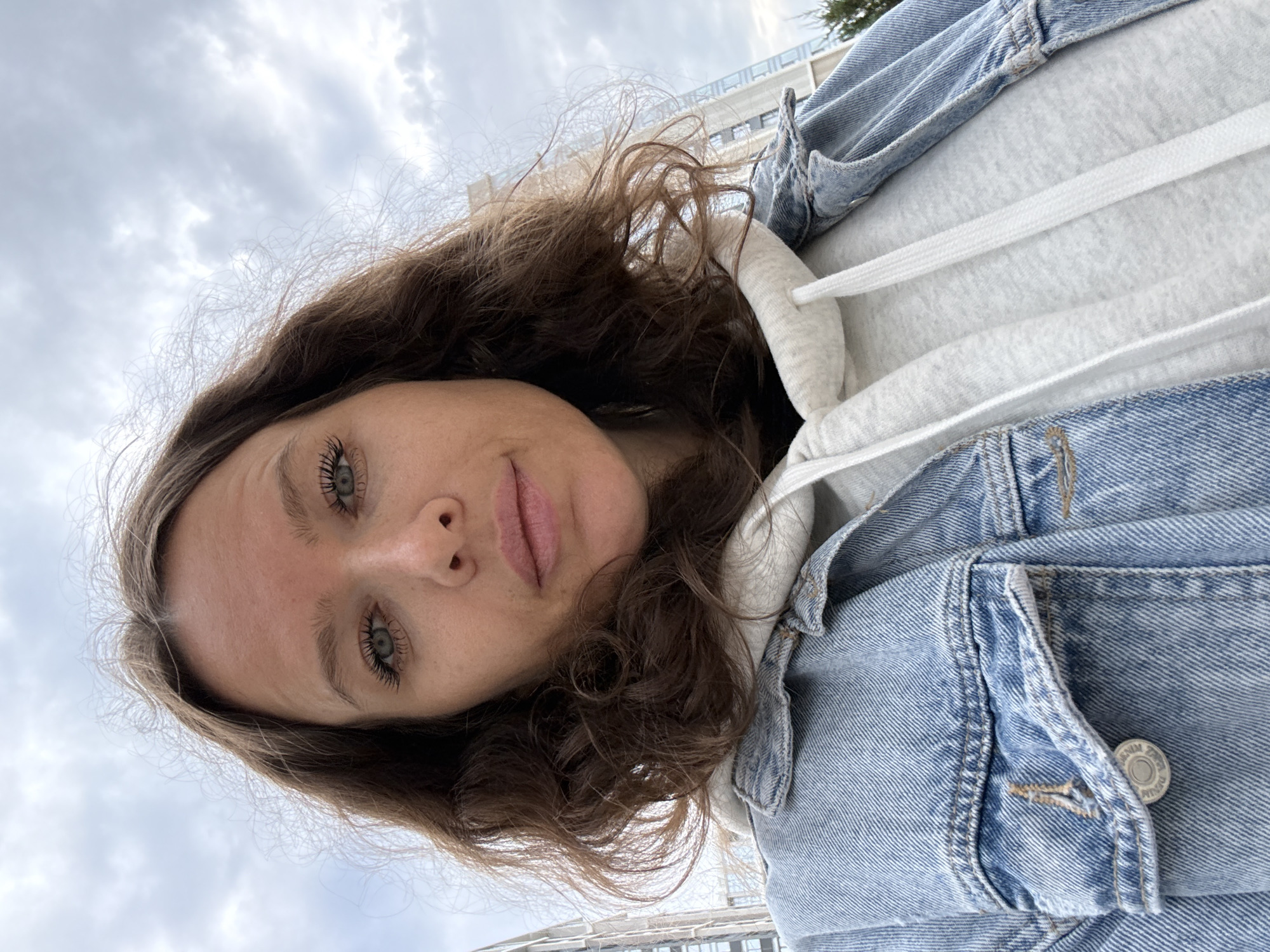 A woman is taking a selfie outdoors. She has brown curly hair and is wearing a denim jacket over a grey hoodie. She has makeup on and her eyes are green. Behind her, tall buildings can be seen against a cloudy sky. The image appears to have been taken in a city.