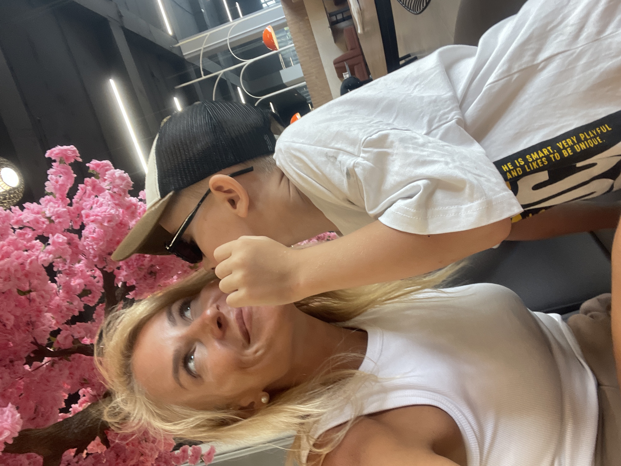 A medium shot captures a blonde woman and a young child in an indoor setting, possibly a cafe or restaurant, adorned with a large artificial pink floral display. The woman, positioned on the left, wears a white tank top and a pearl earring. Her gaze is directed upwards and to the right, with a gentle smile on her face. A young child, on the right, is leaning in towards the woman, whispering in her ear. The child is wearing a white t-shirt with text on it, a baseball cap, and glasses. The background shows interior elements like tables, chairs, and lighting fixtures, with a hint of an outdoor view through large windows. The overall scene evokes a sense of intimate interaction between the two individuals.