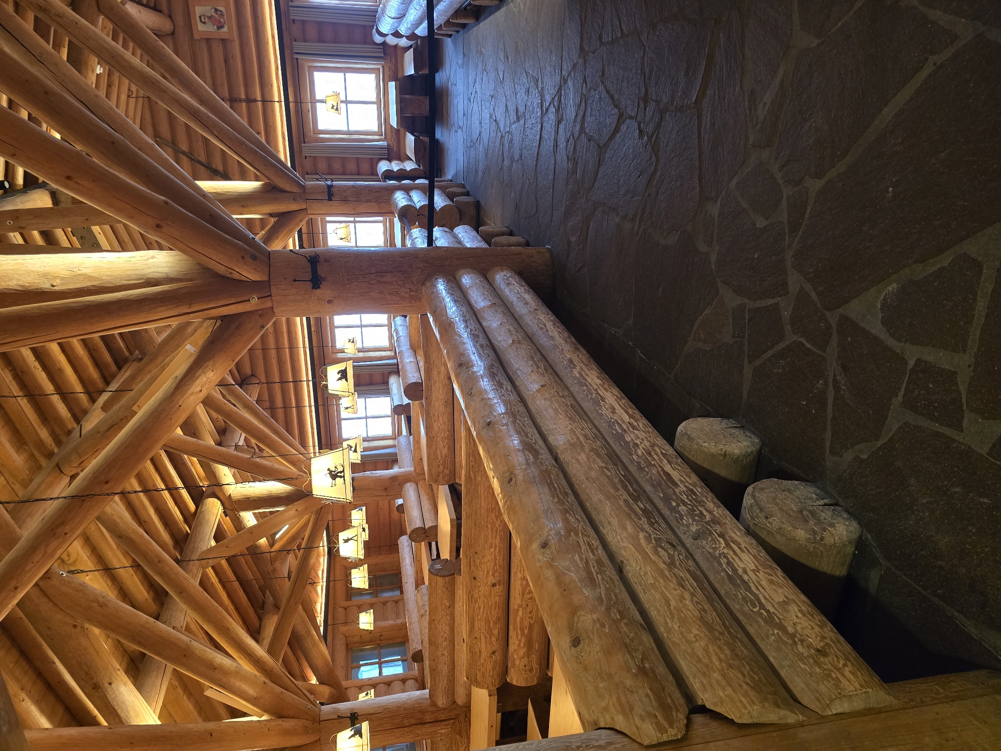 The image depicts the interior of a rustic restaurant with extensive use of natural wood, reminiscent of a mountain lodge. The ceiling features a complex network of exposed wooden beams and log supports, creating a warm and natural aesthetic. Hanging from the beams are several lamps with conical shades, each adorned with silhouette images of skiers. The walls are also constructed from logs, and the furniture consists of sturdy log benches and tables. The floor is paved with irregular, dark-colored stones. Several windows are visible, letting in natural light, and on one wall, a framed portrait is displayed. The overall impression is that of a cozy and traditional dining space, likely in a mountainous region. Given the style, it is plausible this restaurant is located in Garmisch-Partenkirchen, Germany, a town known for its alpine setting and traditional architecture.