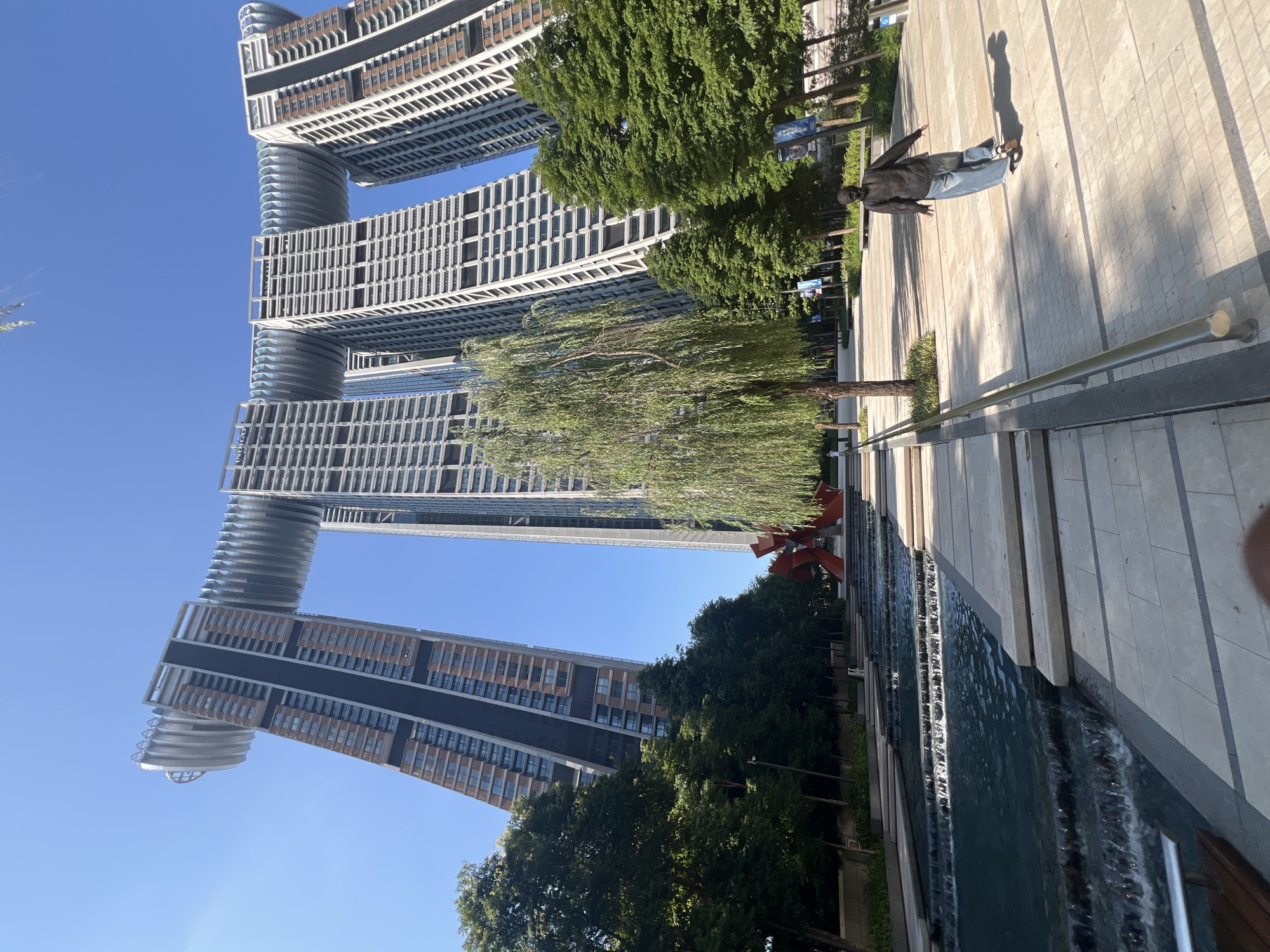 The image captures a low-angle view of the Raffles City Chongqing complex in Yuzhong District, China, on a bright, clear day. The complex consists of multiple skyscrapers that form a horizontal structure across the top, resembling a ship or a dragon. The skyscrapers are modern, with a mix of glass and concrete facades, and some are tilted at an angle. In the foreground, there is a tiered water feature with cascading water and stone steps. A person on a skateboard is moving away from the camera on a paved walkway to the right of the water feature. Lush green trees line the walkway and surround the complex, and a willow tree with drooping branches stands prominently in the middle ground. A large, abstract red sculpture is visible between the water feature and the trees. The sky is a vibrant blue, with no clouds. The overall scene depicts an urban landscape with striking modern architecture, integrated with water features and greenery, and a human presence engaged in recreational activity.