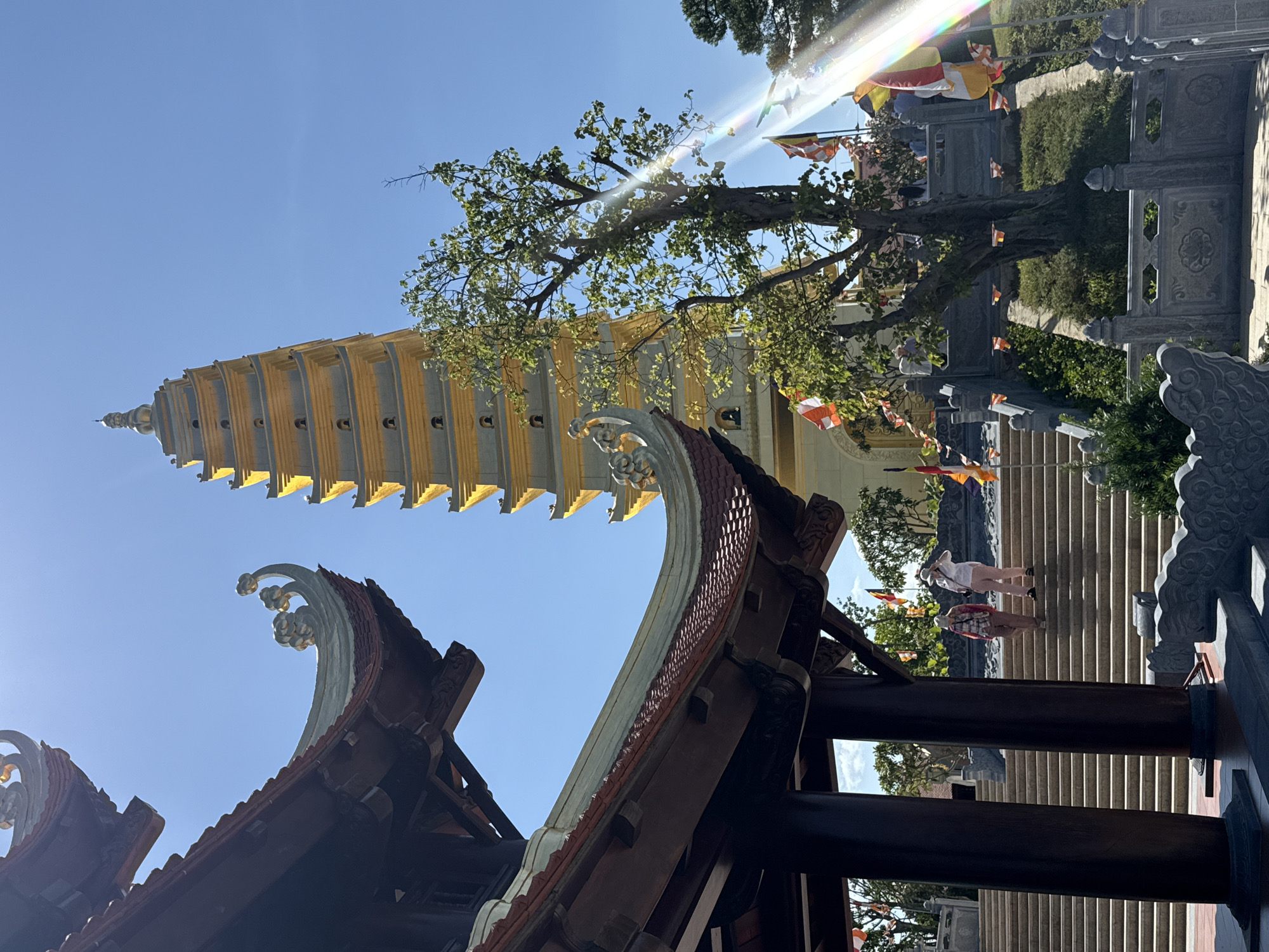 This photograph captures a view of a pagoda in Diên Khánh, Vietnam. The frame is oriented to the side, with a blue sky backdrop. The pagoda, a tall structure with multiple tiers, extends upwards. The architecture shows an Asian style with curved edges. Below the pagoda, there is a building with a traditional, tiled roof. The roof features decorative embellishments, adding to the architectural detail. Steps with two people walking on them are visible behind the pagoda. Flags are seen at the top of the steps with some trees. The weather looks sunny, there are rays of light visible.