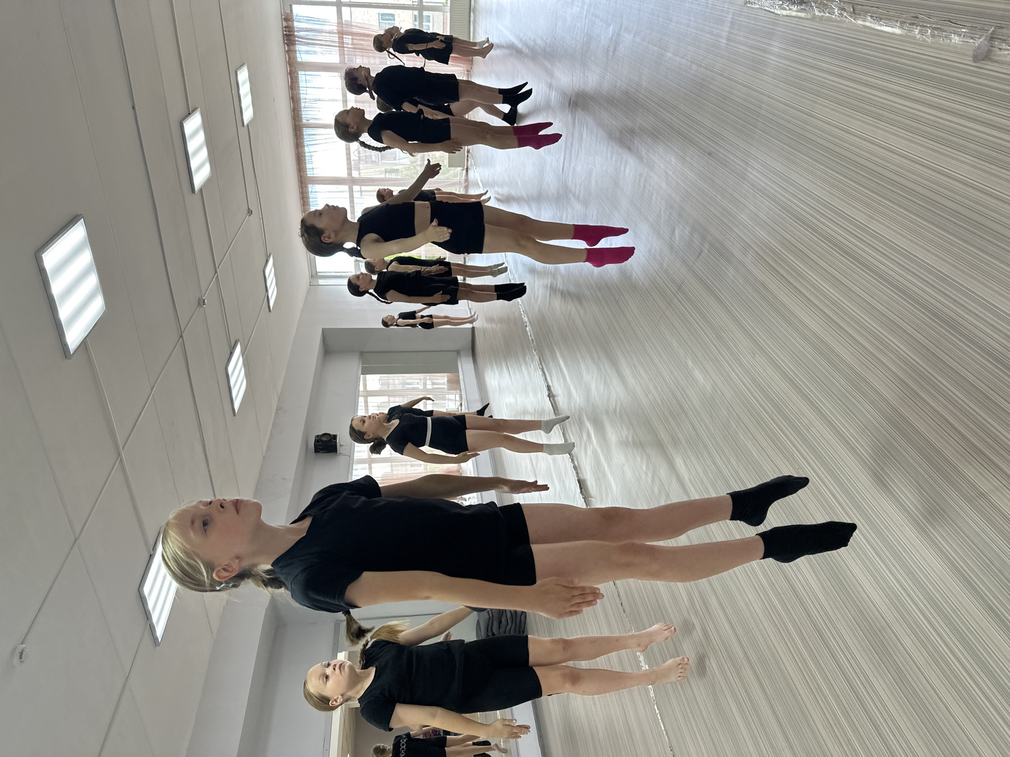 A group of approximately 10 young girls are lying in a row on a light-colored floor. They are oriented in a line from bottom-left to upper-right. The girls are wearing black leotards or short dresses. Some are wearing socks of various colors, including black, white, and pink. The girls are facing upwards and have their arms extended to the sides, forming a "T" shape with their bodies.
The setting appears to be a dance or exercise studio. A row of windows can be seen in the upper part of the image, along with multiple rectangular ceiling lights. Based on the visible buildings and window placement, along with the general architectural style and presence of dance studios in educational facilities, the location is possibly a dance studio or gymnastics hall within a school or community center in Votkinsk, Russia. The floor has a wood-grain or striped pattern. The room appears to be well-lit and organized, possibly for a dance or rhythmic gymnastics class.