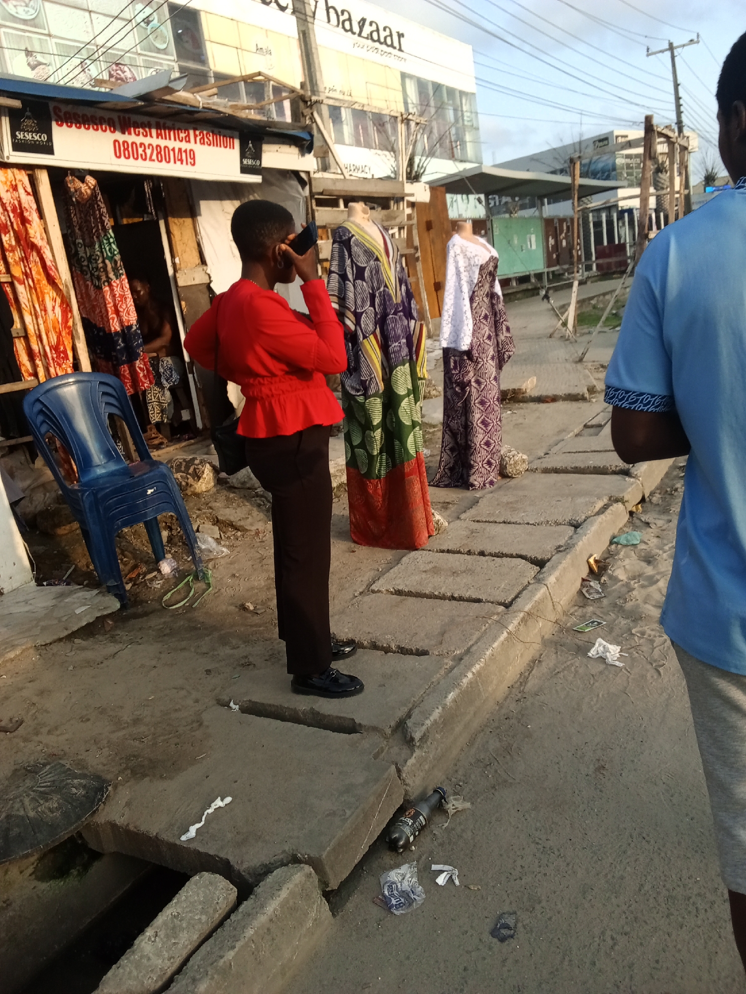The image depicts a street scene in Ado, Nigeria, with "Sosesco West Africa Fashion" visible on the side of a building. A woman in a red top and black trousers is talking on a cell phone. Clothing is displayed on mannequins outside a store. A blue plastic chair is stacked with other chairs. Debris including a plastic bottle, wrappers, and paper are present on the ground. A man in a blue shirt is partially visible on the right side of the image.